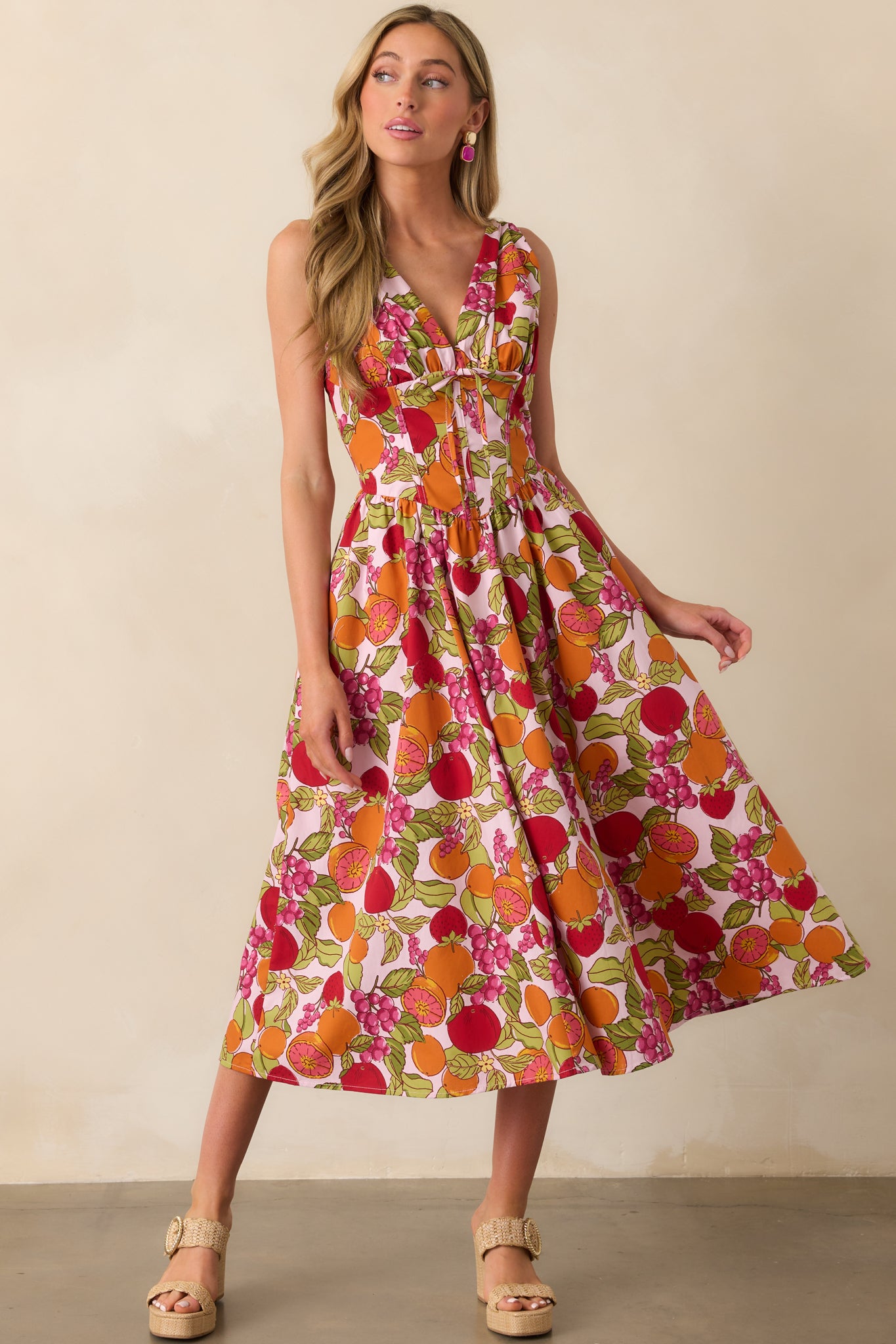 Front view of the pink maxi dress featuring a multicolor fruit print, deep v-neckline, and a decorative bow at the center bust.