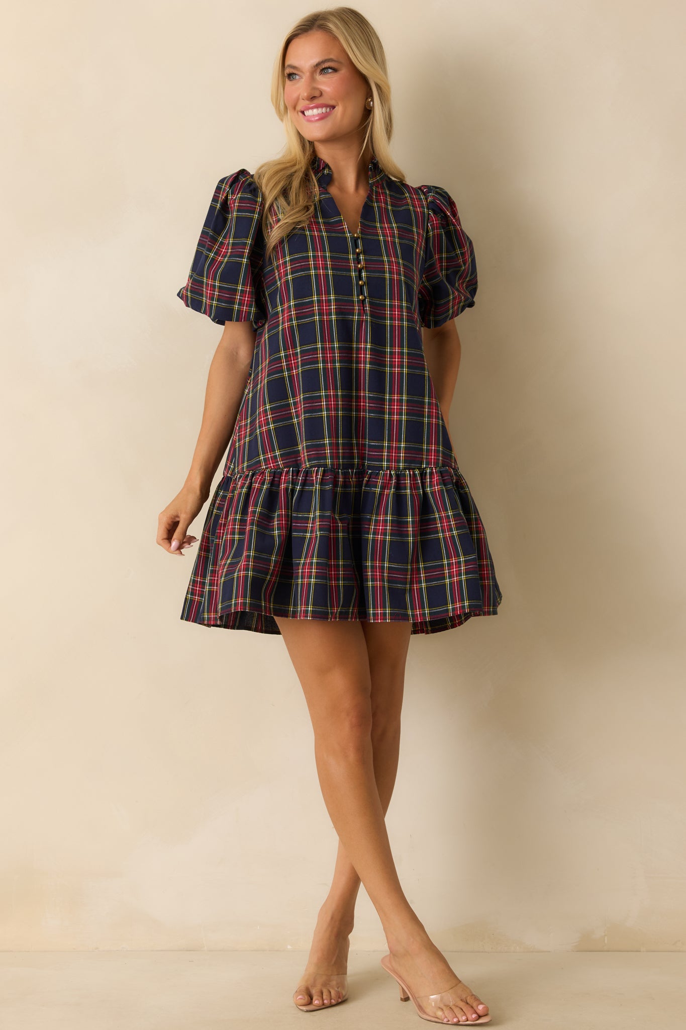 Full-length front view of navy plaid mini dress featuring short puff sleeves, split neckline with button detail, side pockets, and ruffled drop hem.