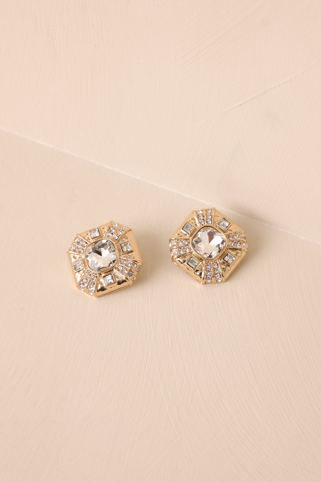 The gold and rhinestone earrings displayed against a neutral beige background, showcasing their full shape and elegant design.