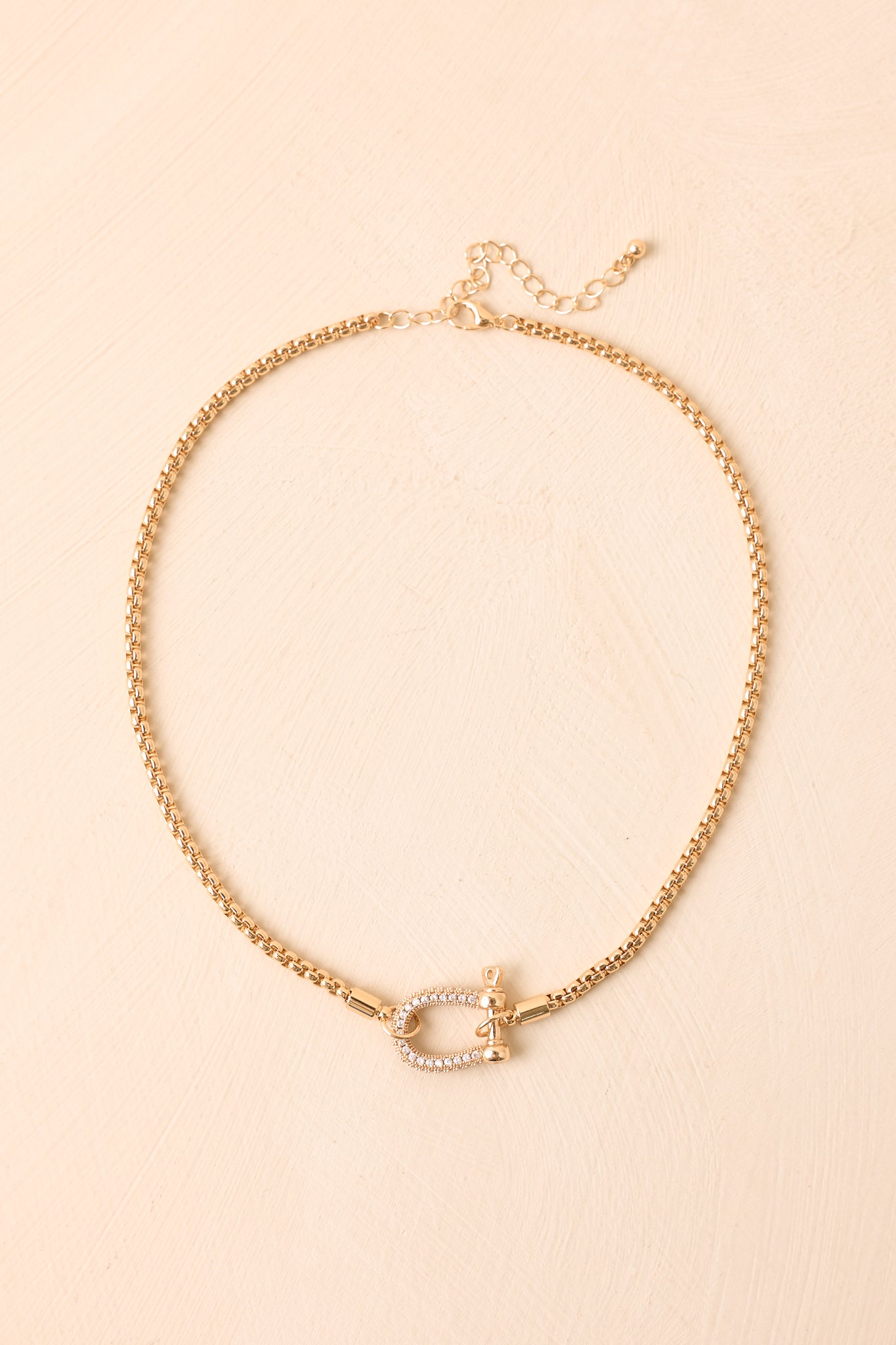 Gold-tone necklace displayed against a neutral background, showcasing the structured box chain and sparkling clasp centerpiece.