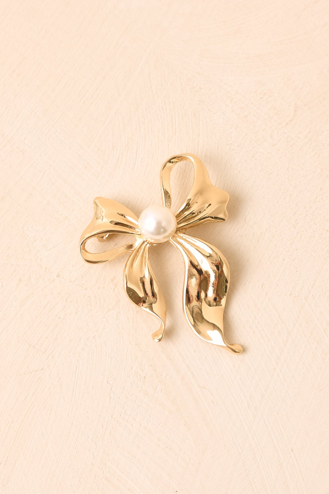 Close-up of the gold-tone brooch showcasing the luminous pearl center, layered ribbon texture, and refined sculptural craftsmanship.