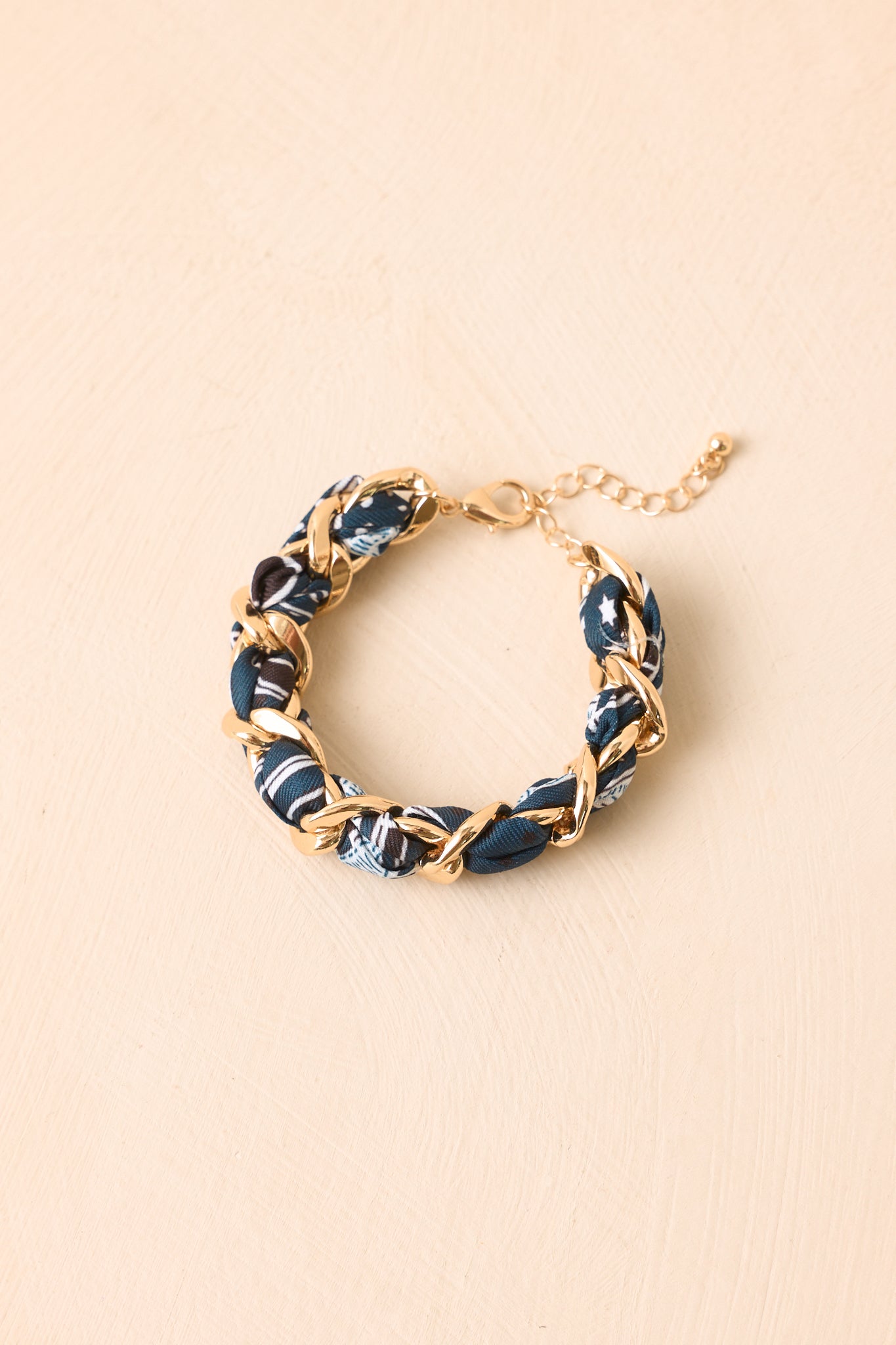 A gold chain bracelet with alternating links of dark blue and white patterned fabric, displayed on a light beige textured background.