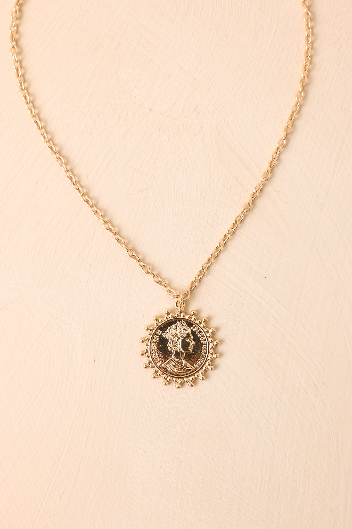 Gold coin pendant necklace displayed against a beige background, highlighting the detailed charm, beaded frame, and polished chain.