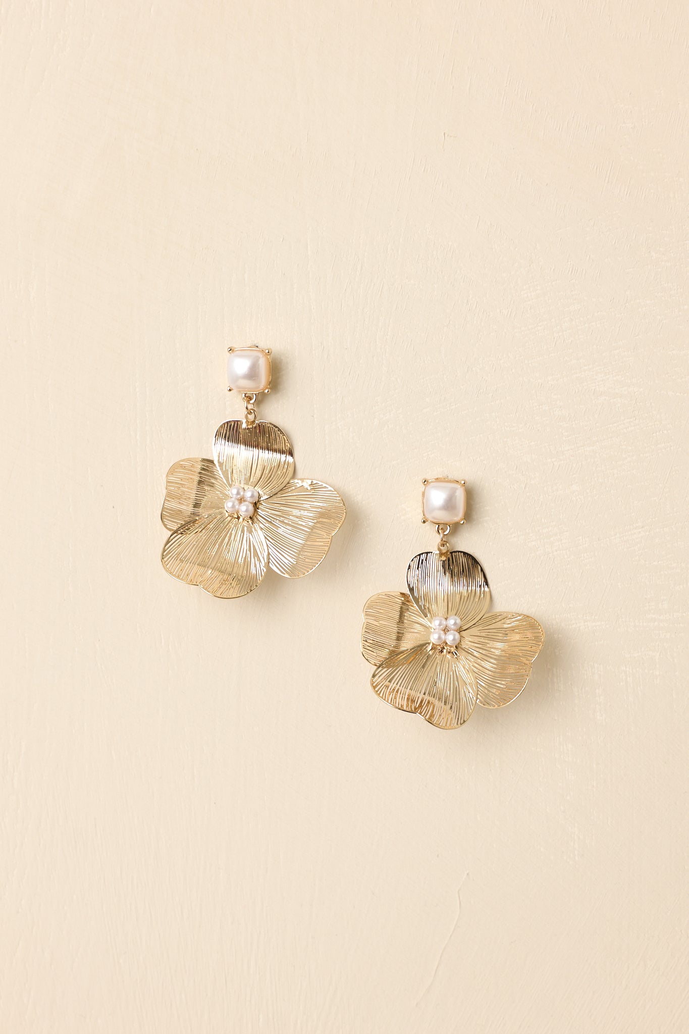 Gold floral drop earrings displayed on a beige background, highlighting textured petals and luminous pearl cluster.