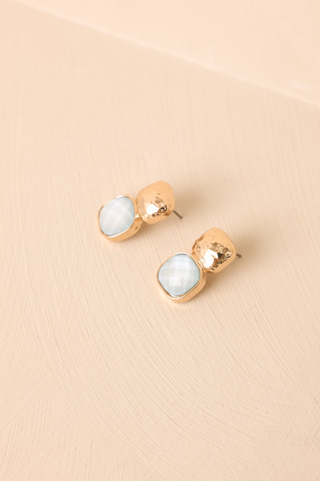 Gold hammered drop earrings displayed against a beige background with ice blue stone accents.