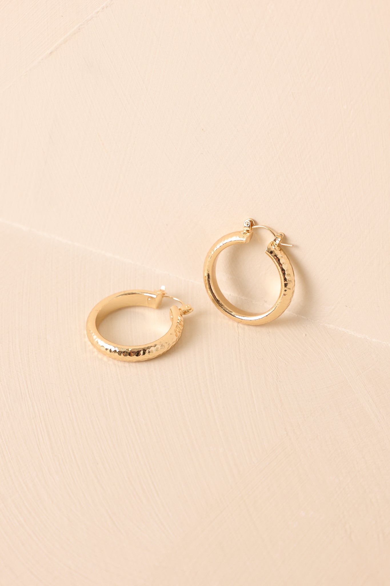 Gold hammered earrings displayed against a beige background, emphasizing the organic texture and curved shape.