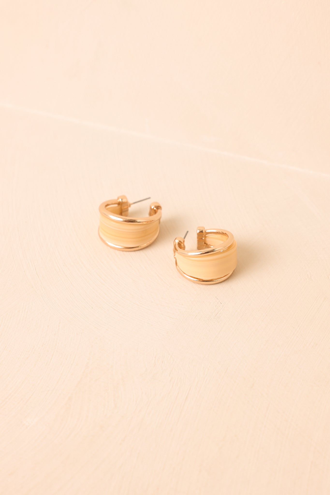 Polished gold-tone hoops with an ivory resin inset presented on a neutral beige backdrop for an elegant, contemporary look.