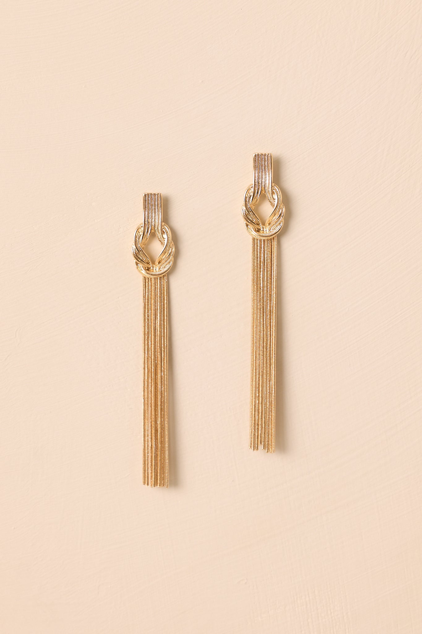 Gold-tone knot tassel earrings displayed against a beige background, showcasing the cascading chain design.