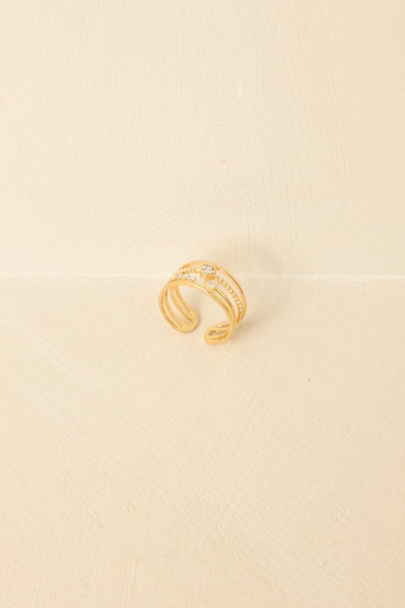 Gold layered ring displayed on a beige background, highlighting slender bands and subtle rhinestone detailing.