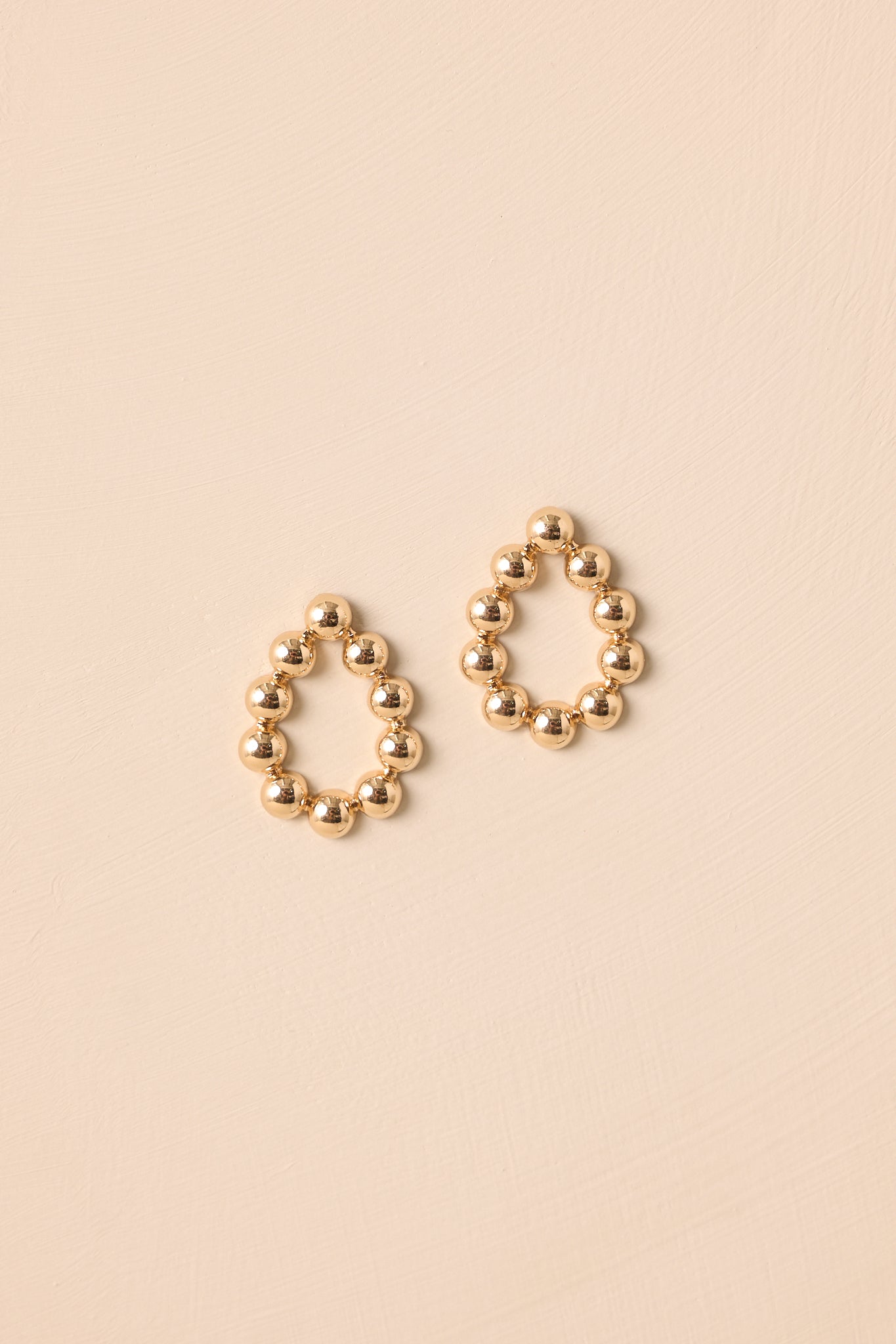 Gold open teardrop earrings displayed against a beige background, featuring the consistent circular bead pattern.