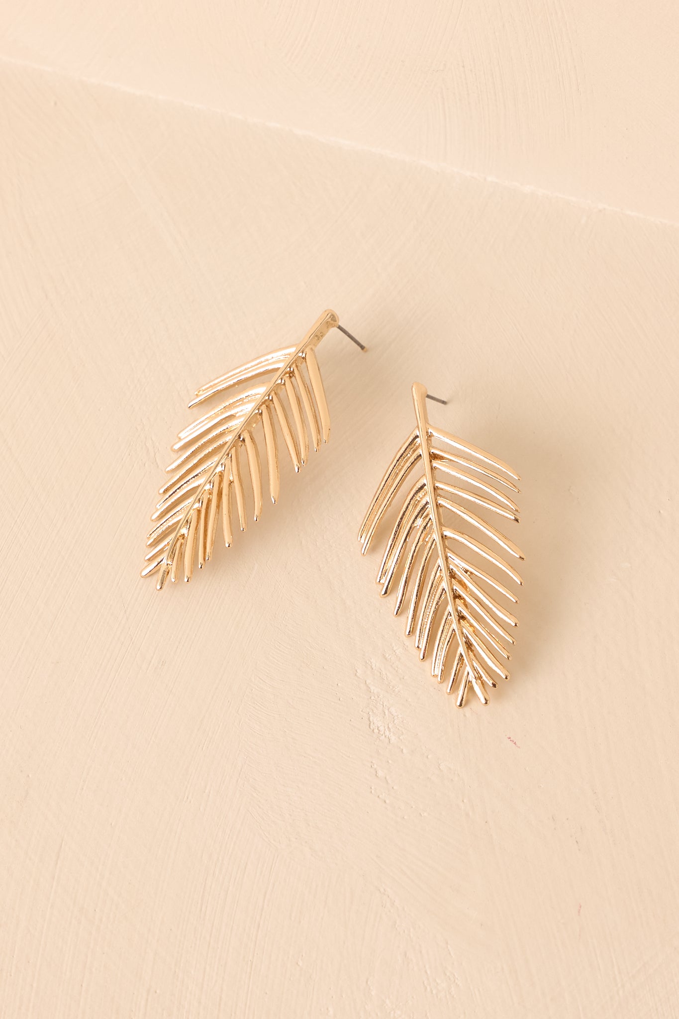A shot of the gold palm leaf drop earrings with a curved, textured design displayed against a beige background.