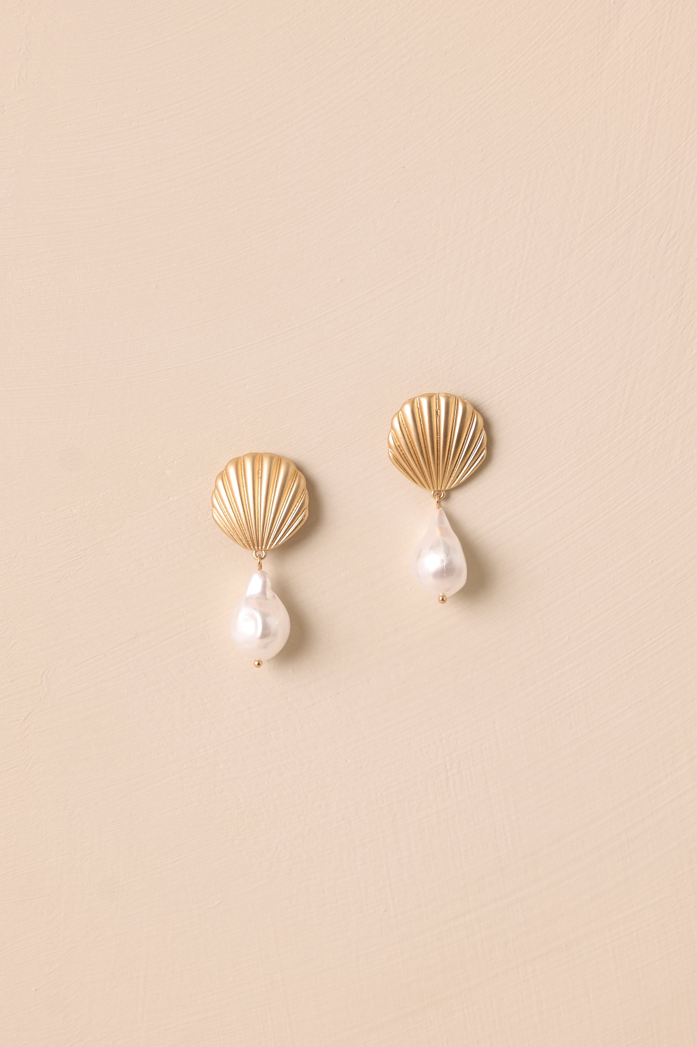 Gold pearl drop earrings displayed against a beige background featuring seashell studs and textured details.
