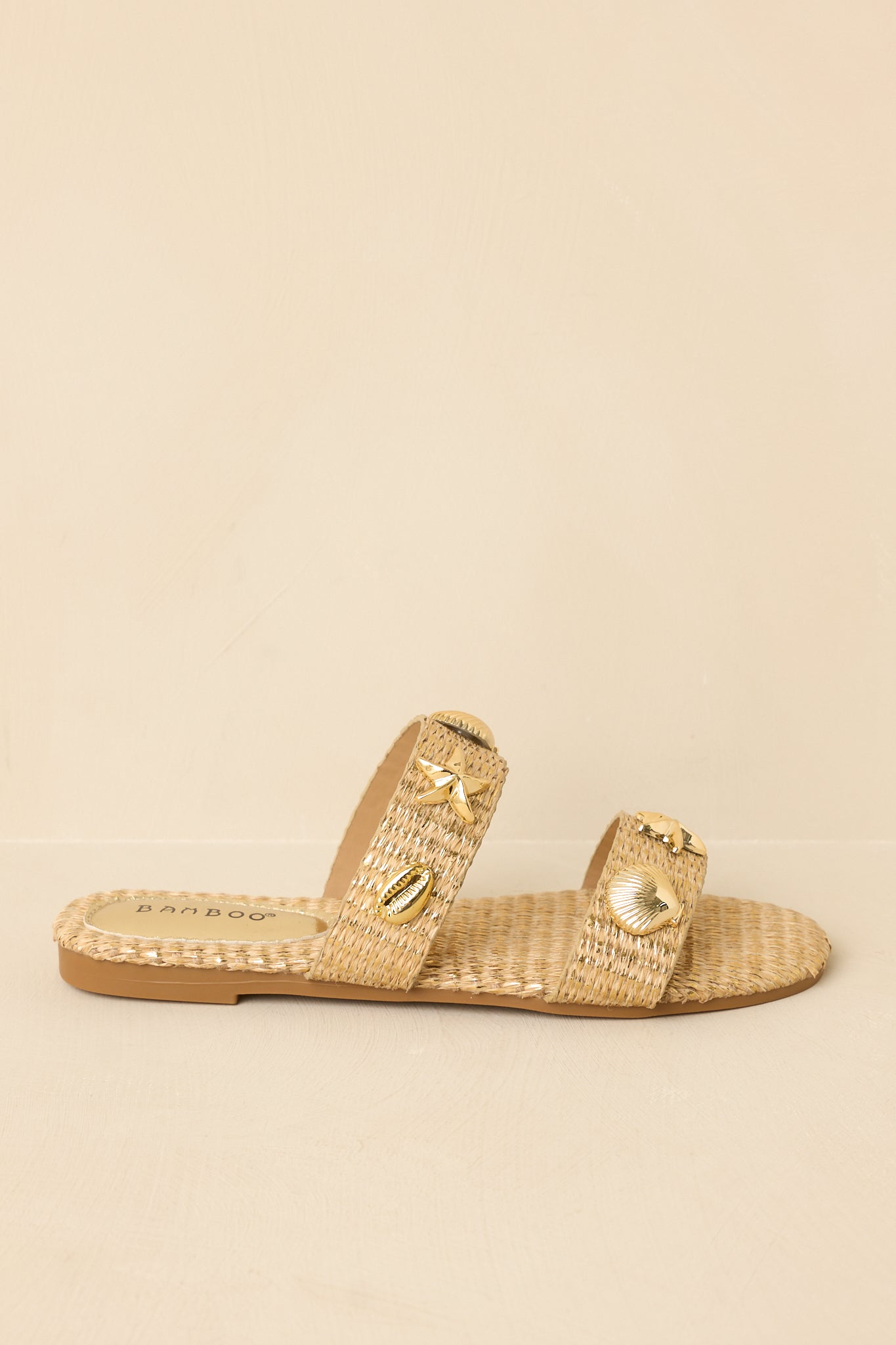 Side view of gold raffia slip-on sandals on a beige background highlighting the woven texture and flat sole.