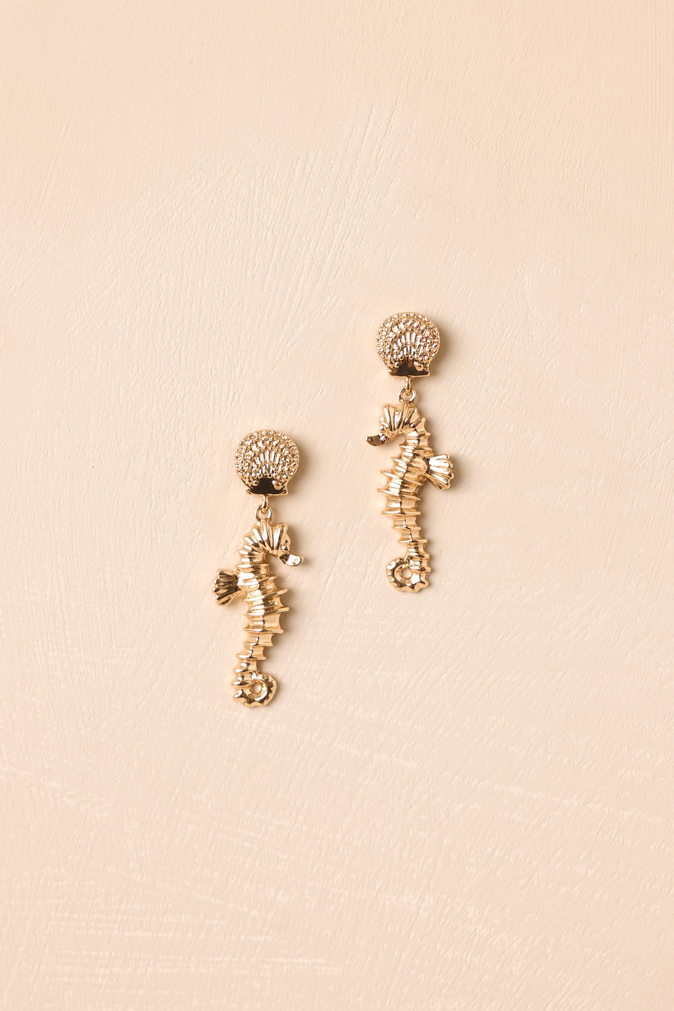 Gold seahorse drop earrings displayed on a beige background emphasizing the textured shell stud and polished gold finish.