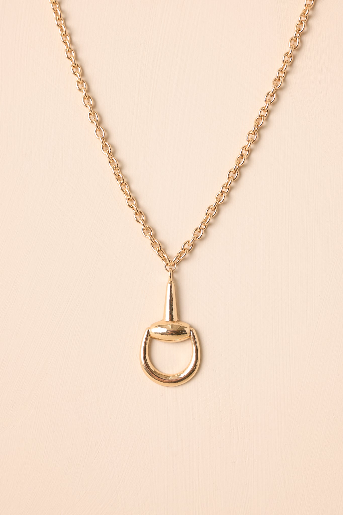 Gold stirrup pendant necklace displayed on a beige background, showing the chain and lobster clasp closure.