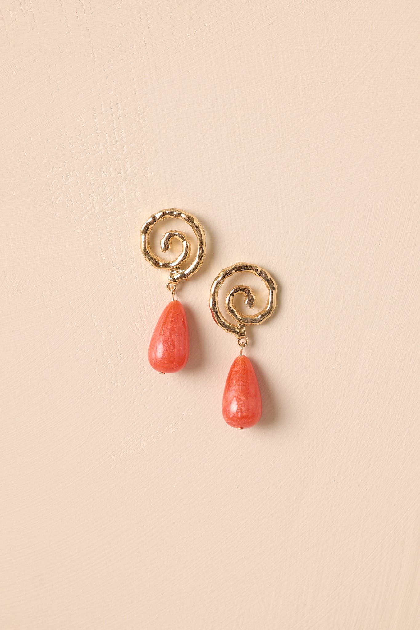 Gold swirl stud earrings with pearlescent terracotta drops displayed on beige backdrop.
