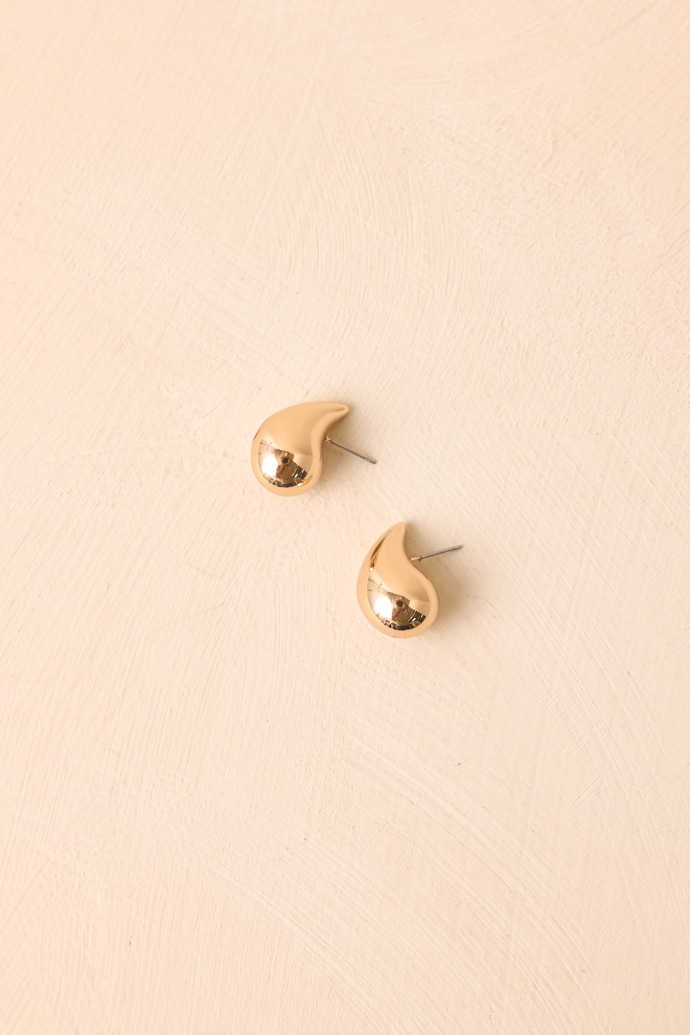 Gold teardrop earrings displayed against a beige background, featuring a sleek sculptural shape and smooth high-shine finish.