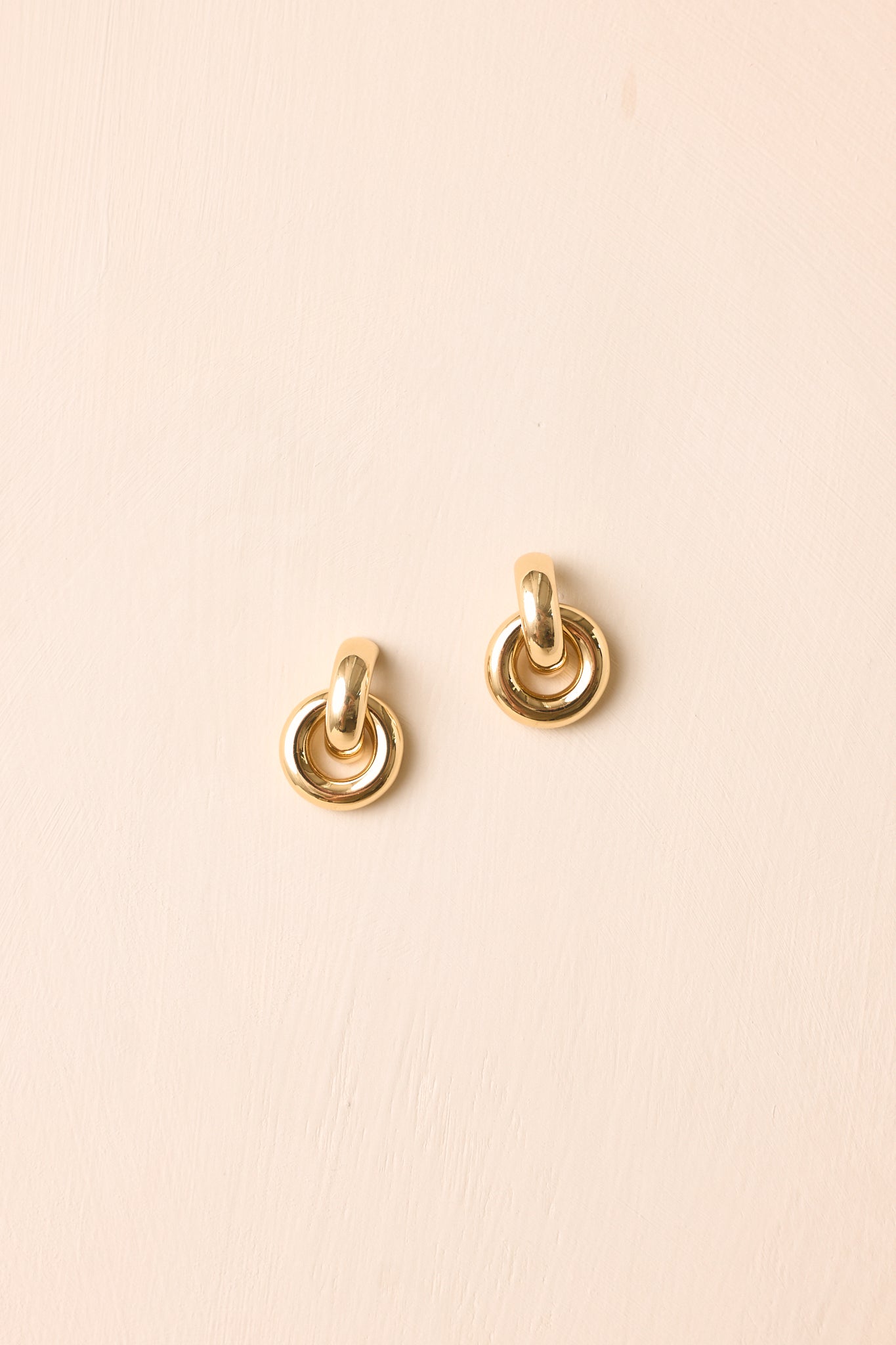 Gold-tone earrings displayed against a beige background, featuring an interlocking ring design with a smooth rounded silhouette and polished finish.