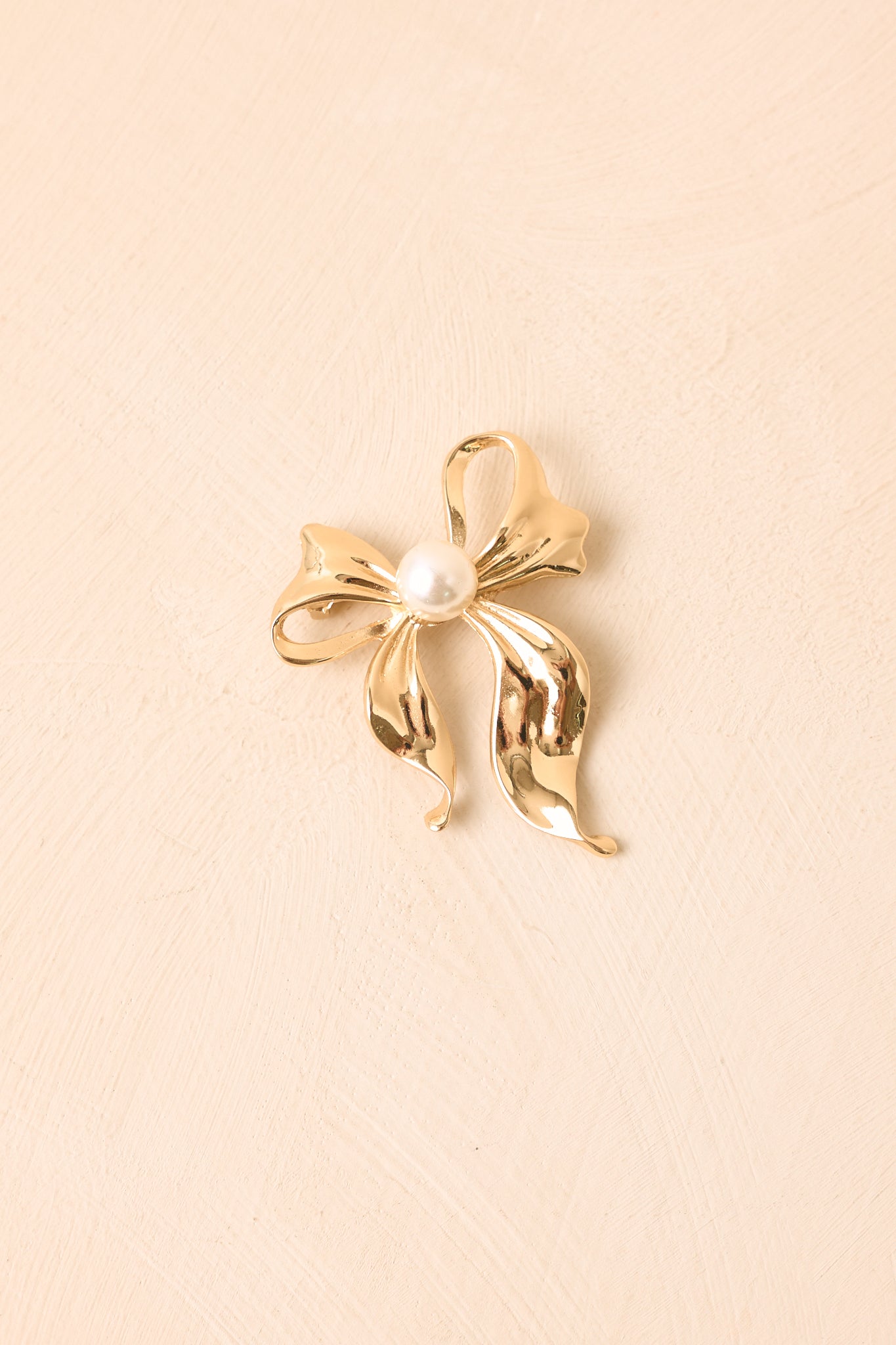 Gold-tone ribbon-inspired brooch featuring softly curved loops, flowing tails, and a luminous pearl center with a sculptural bow silhouette.