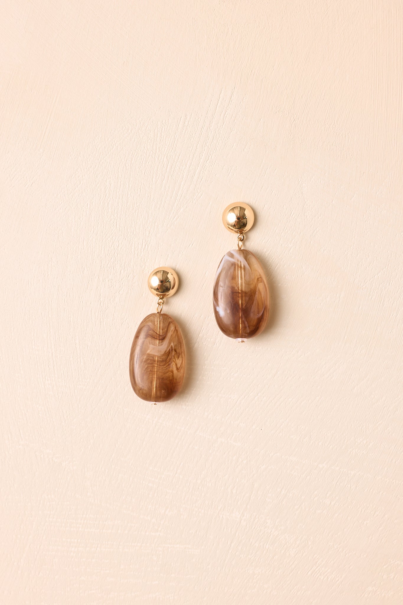 Gold-tone stud earrings featuring smooth marbled tortoise beads, elongated oval shapes, and a polished finish with a modern statement silhouette against a beige background.