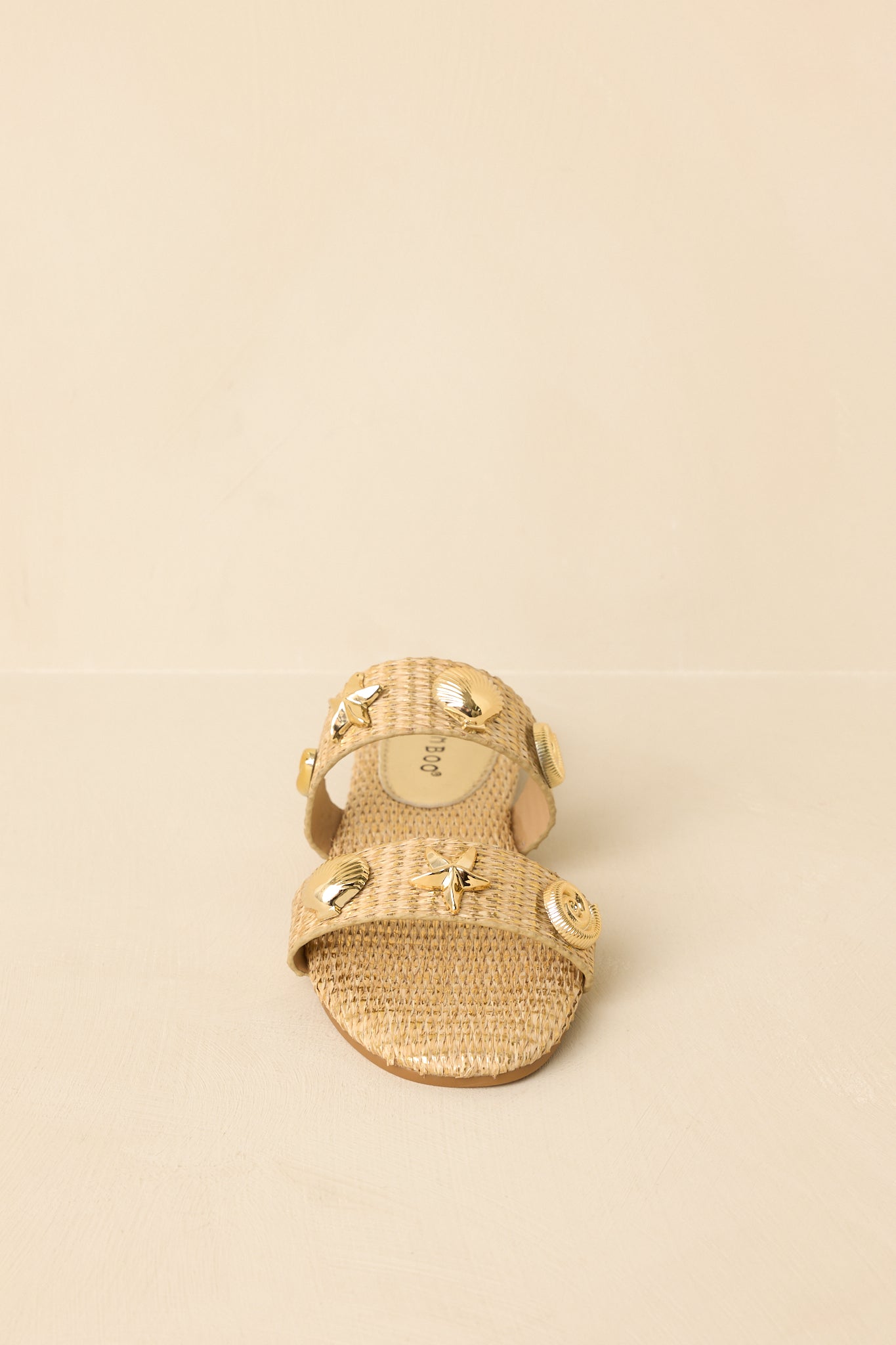 Front view of gold woven sandals on a beige background emphasizing the open toe and embellished straps.