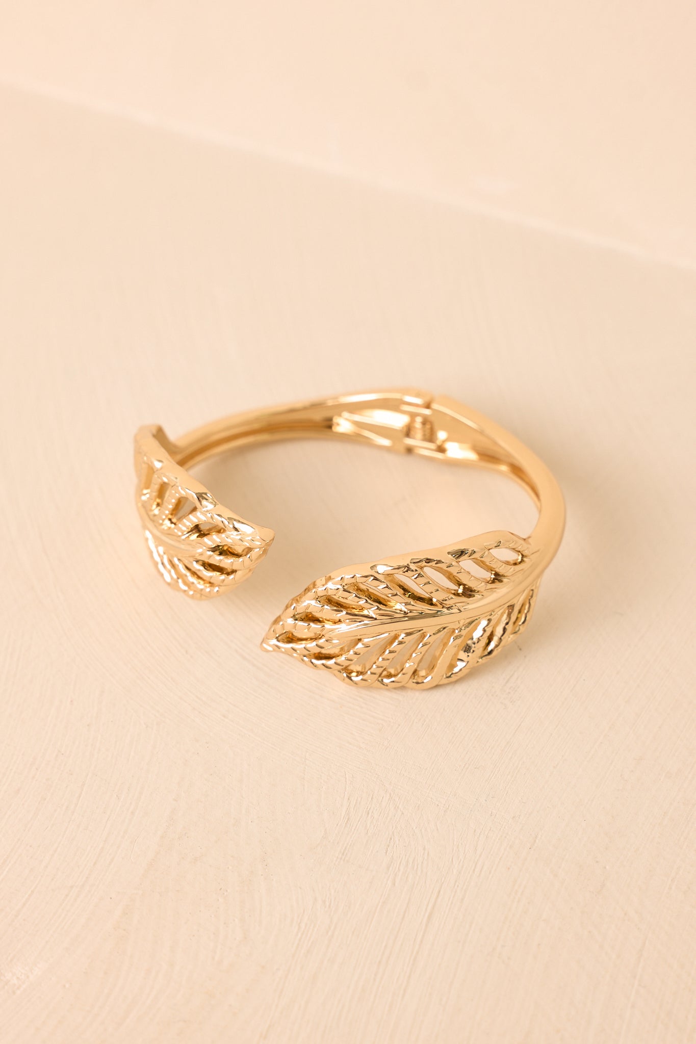 Gold wrap bracelet showcasing intricate leaf detailing and a hinged closure displayed against a beige background.