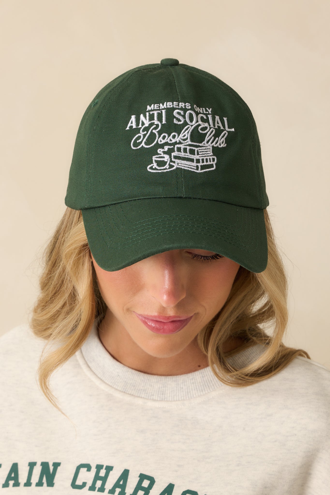 A cropped front view of the green cotton twill baseball cap with a curved brim and raised white embroidered “Anti Social Book Club” graphic.