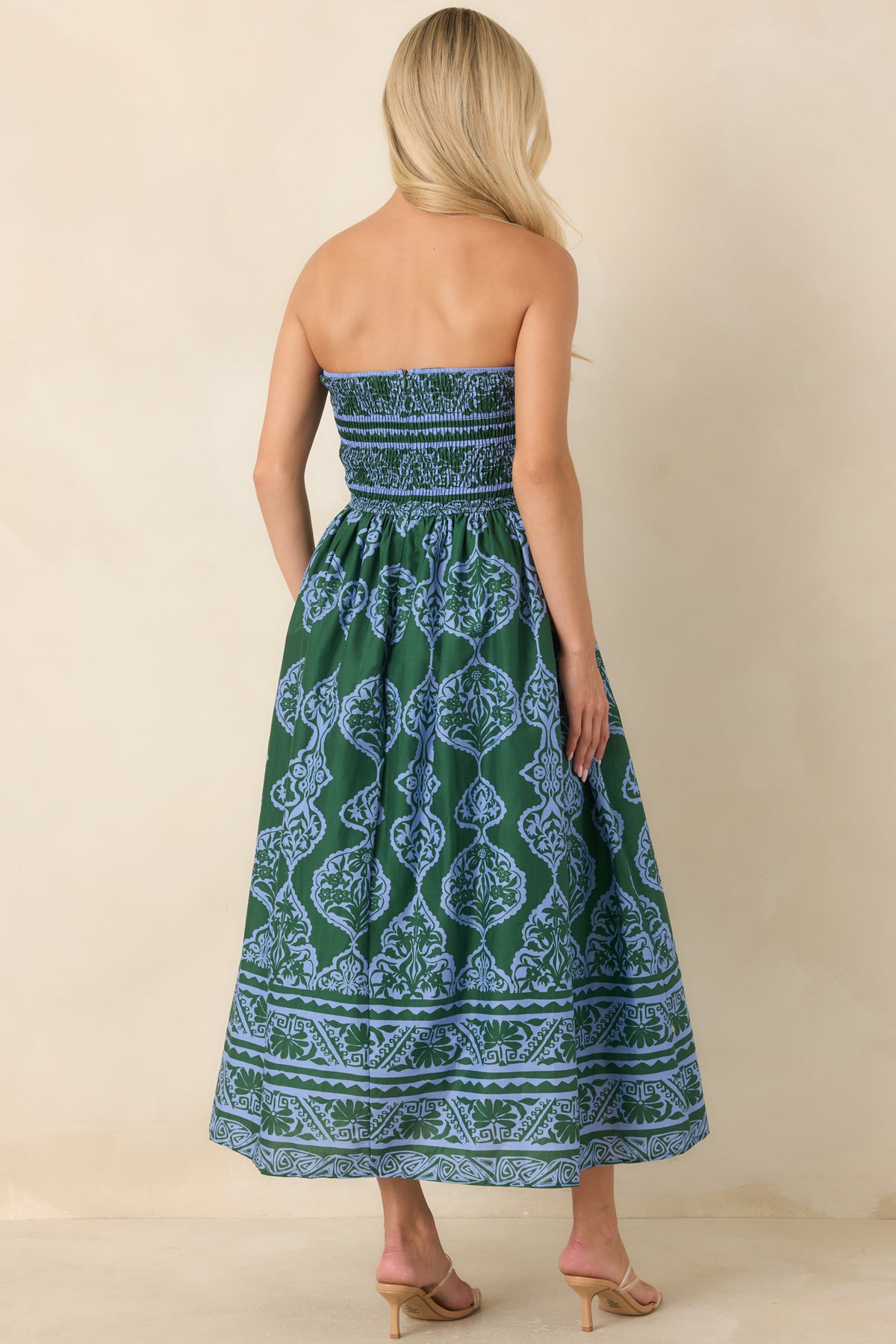 A woman shown from the back wearing a strapless maxi dress with a green and blue paisley pattern, standing against a plain beige studio backdrop.