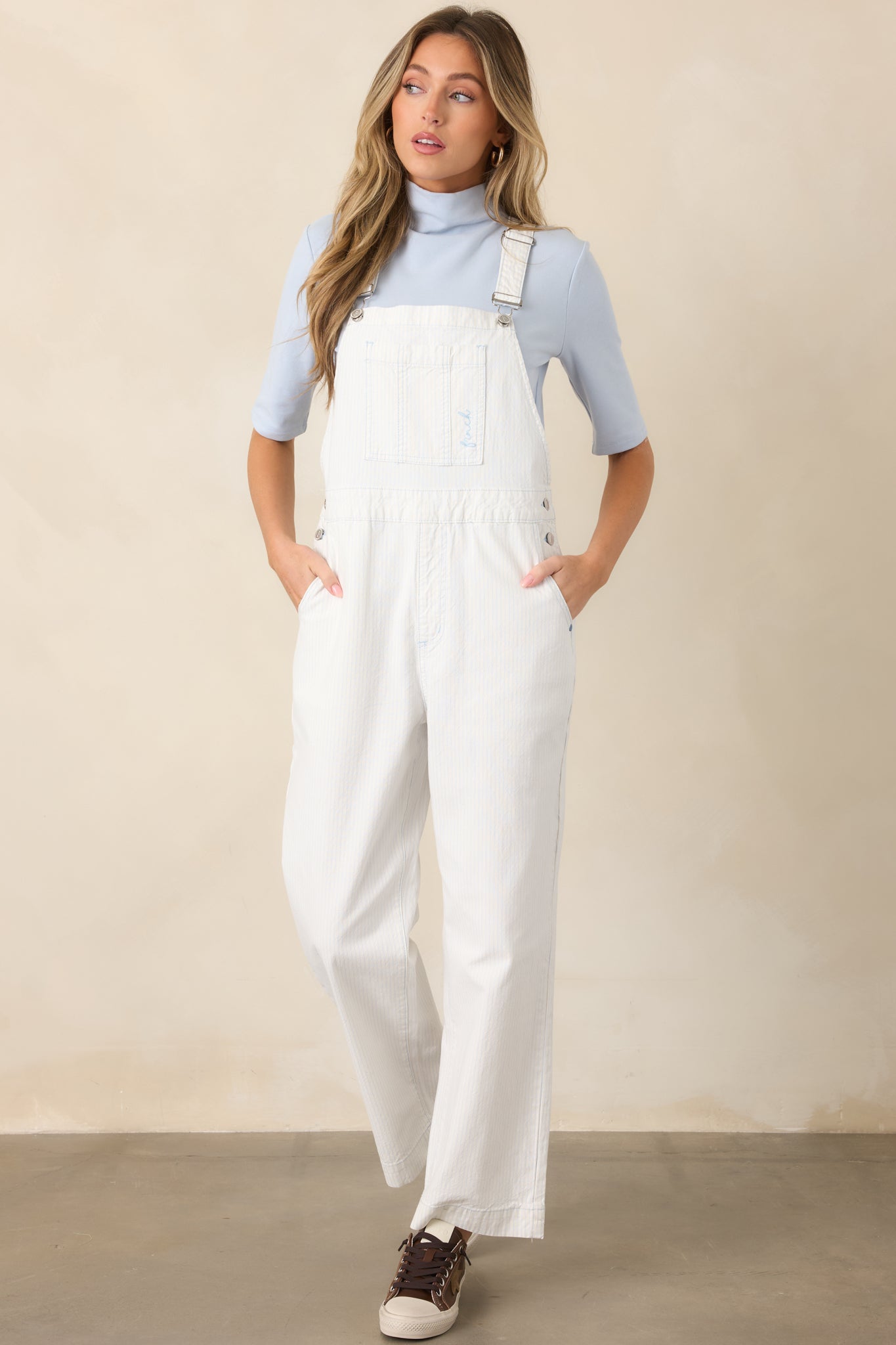 Ivory and sky blue striped cotton overalls with adjustable buckle straps, a relaxed straight-leg fit, chest pocket, side button accents, and ankle-length hems.