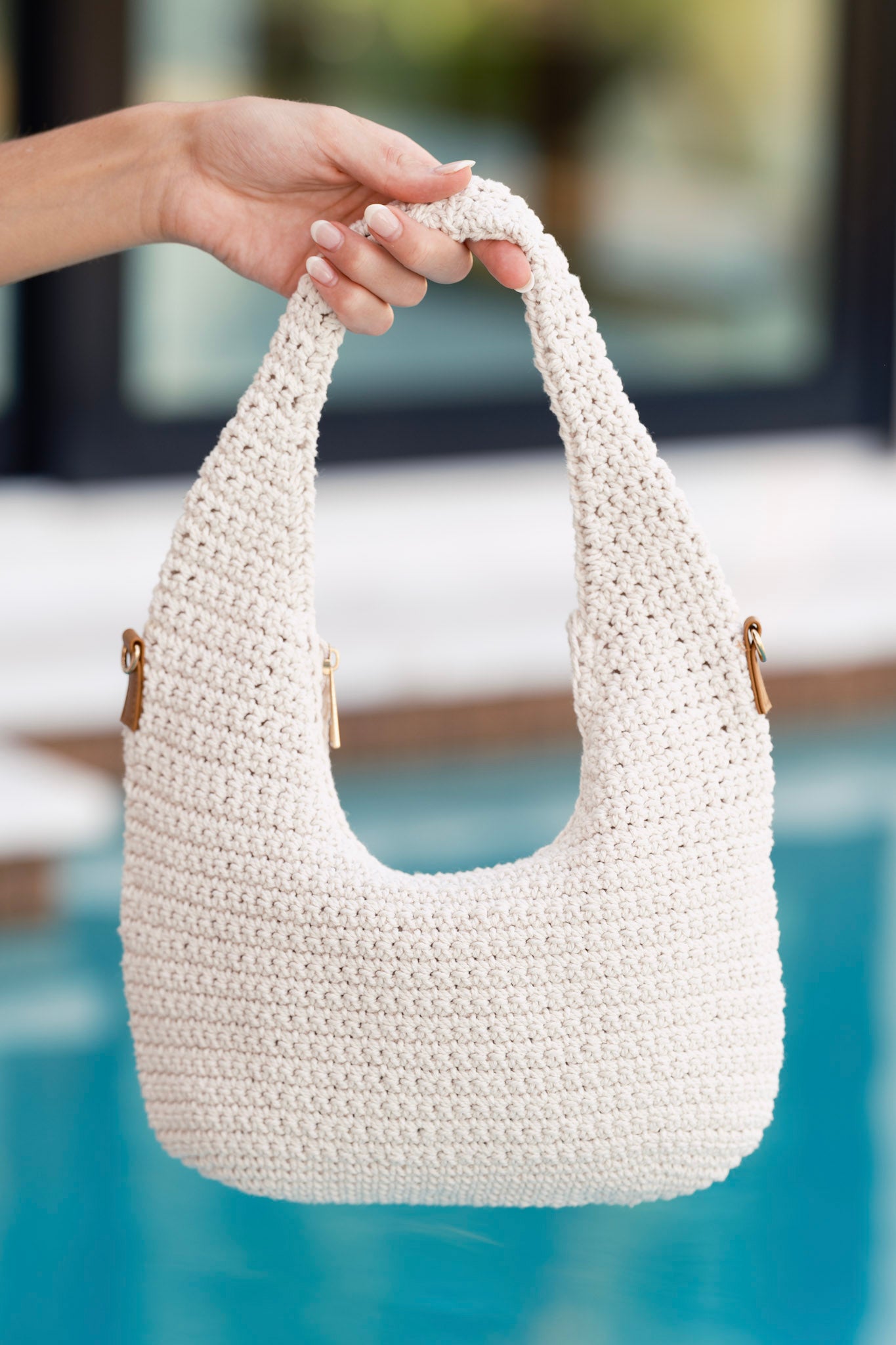 Ivory handbag with woven crochet fabric, zipper closure, and optional gold crossbody chain, shown held by the handle over a pool background.