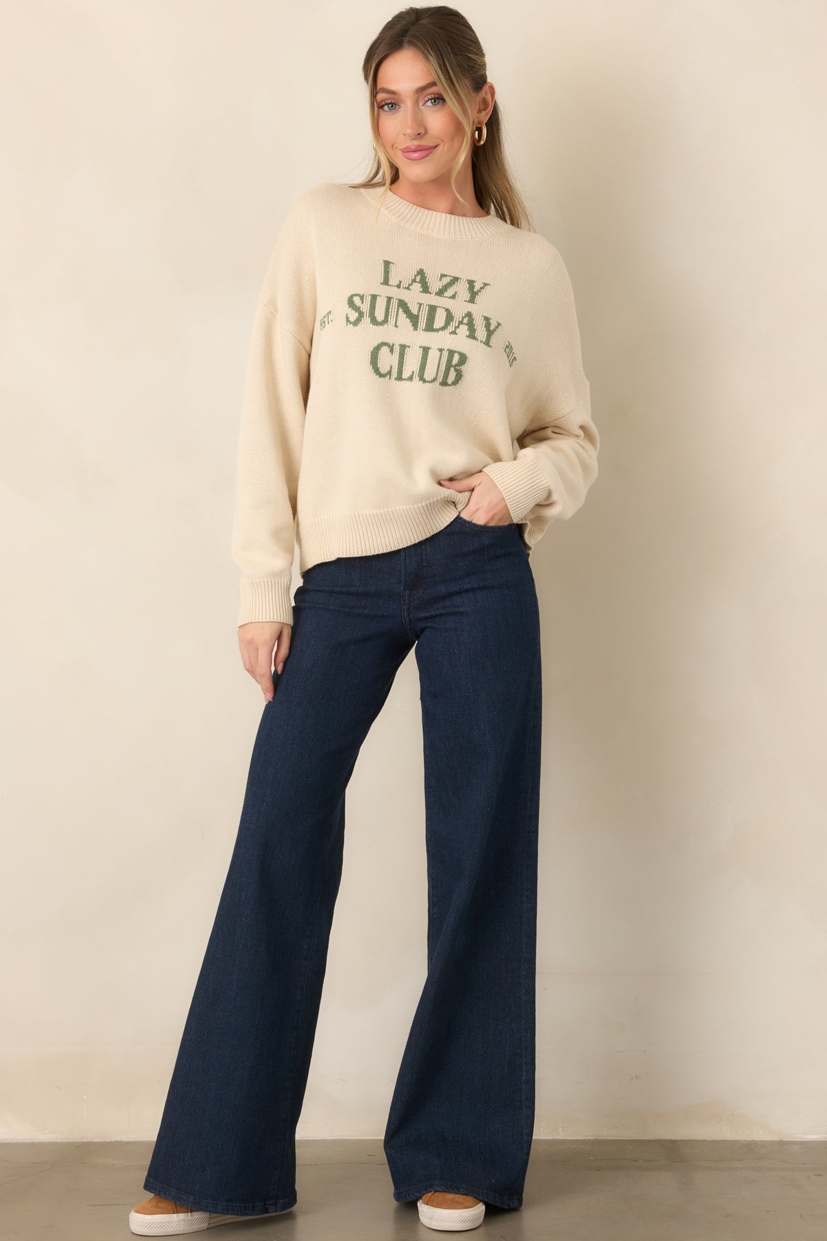 Ivory knit pullover sweater featuring a crew neckline, long drop-shoulder sleeves, ribbed hems, and green “LAZY SUNDAY CLUB” chest graphic.
