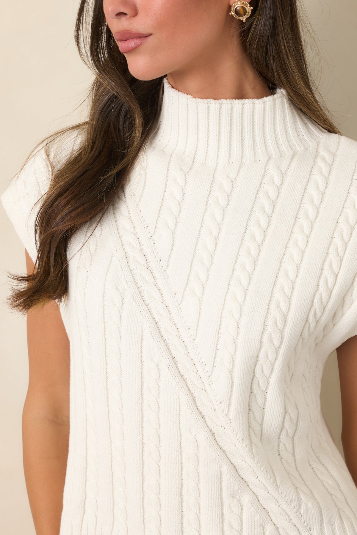 The ivory sweater’s relaxed sleeveless shape continues into a clean ribbed hem beneath the structured cable-knit pattern.