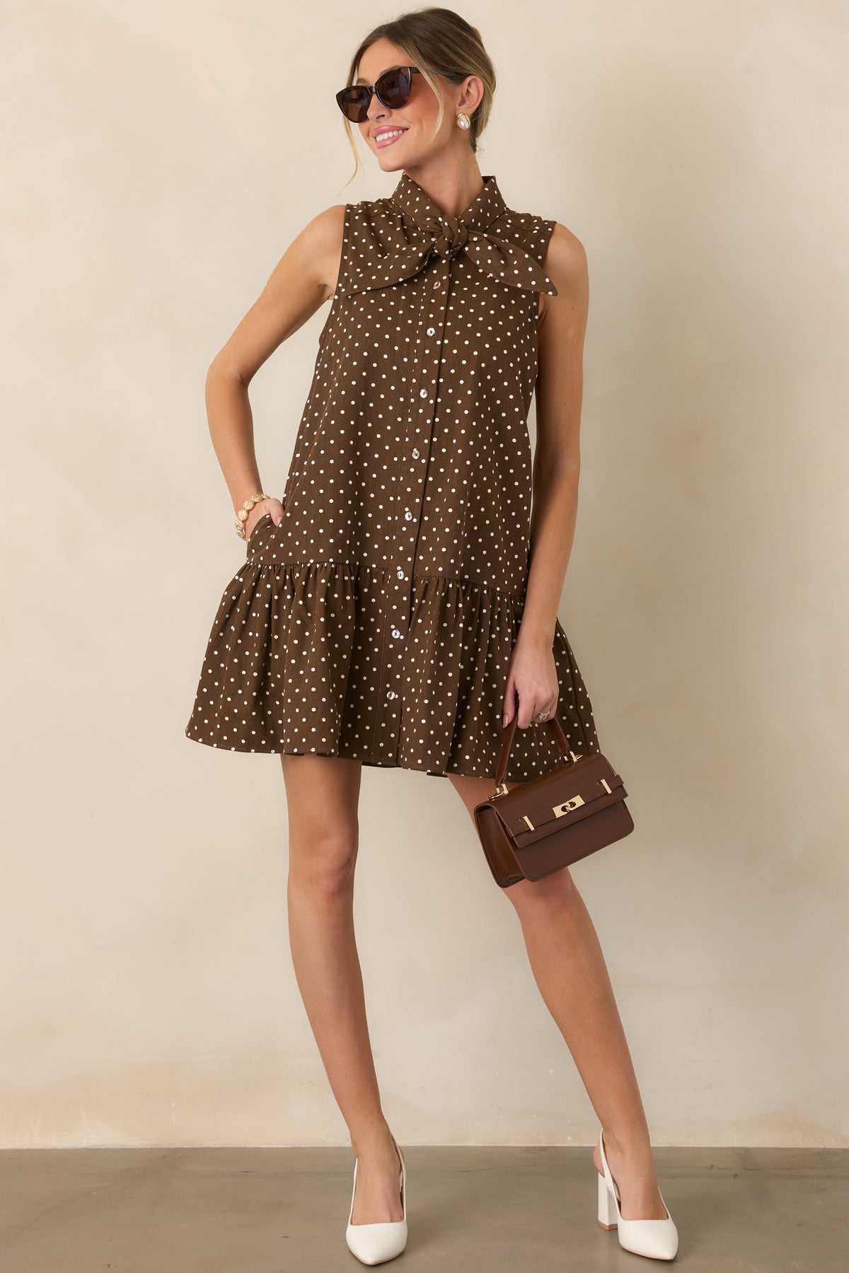Sleeveless ivory polka dot dress featuring a relaxed drop-waist silhouette, adjustable self-tie neckline, button-front design, and voluminous ruffle hem.