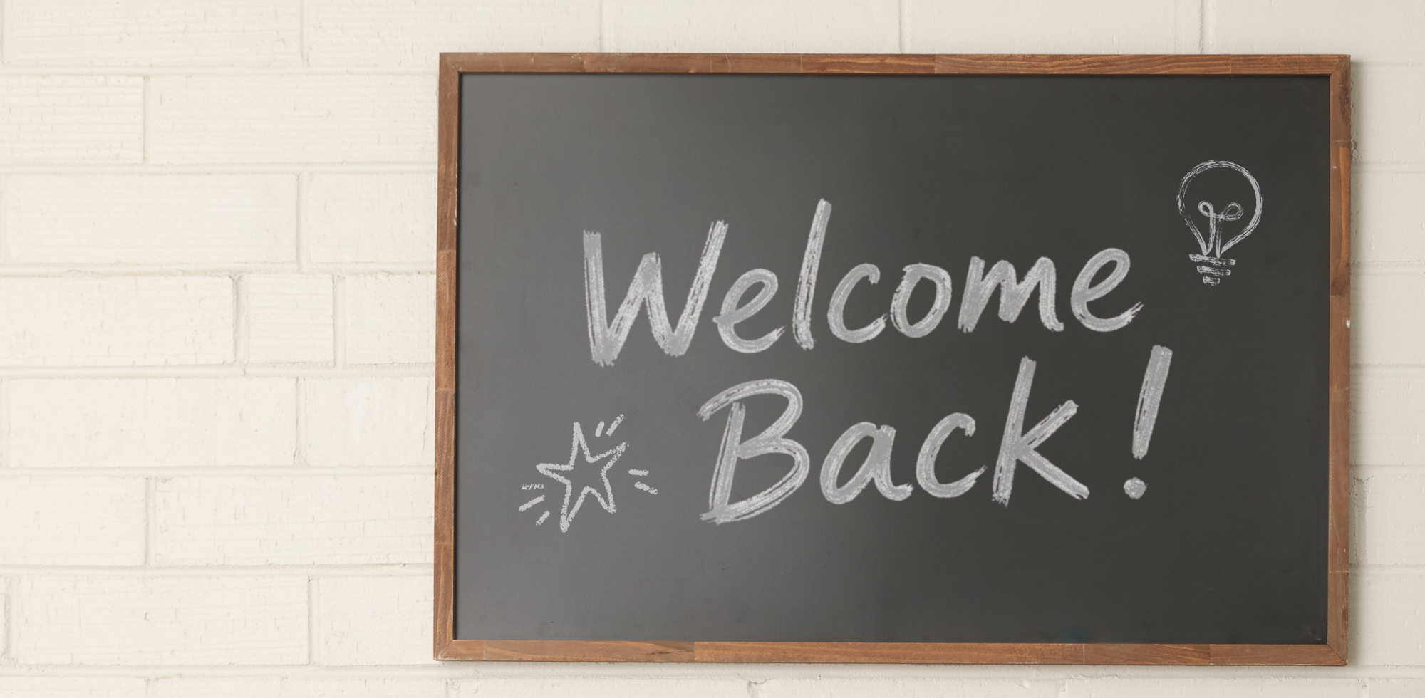 A rectangular black chalkboard with a wooden frame hangs on a white painted brick wall. Handwritten white chalk text on the board reads 'Welcome Back!' with small chalk drawings of a star and a lightbulb.