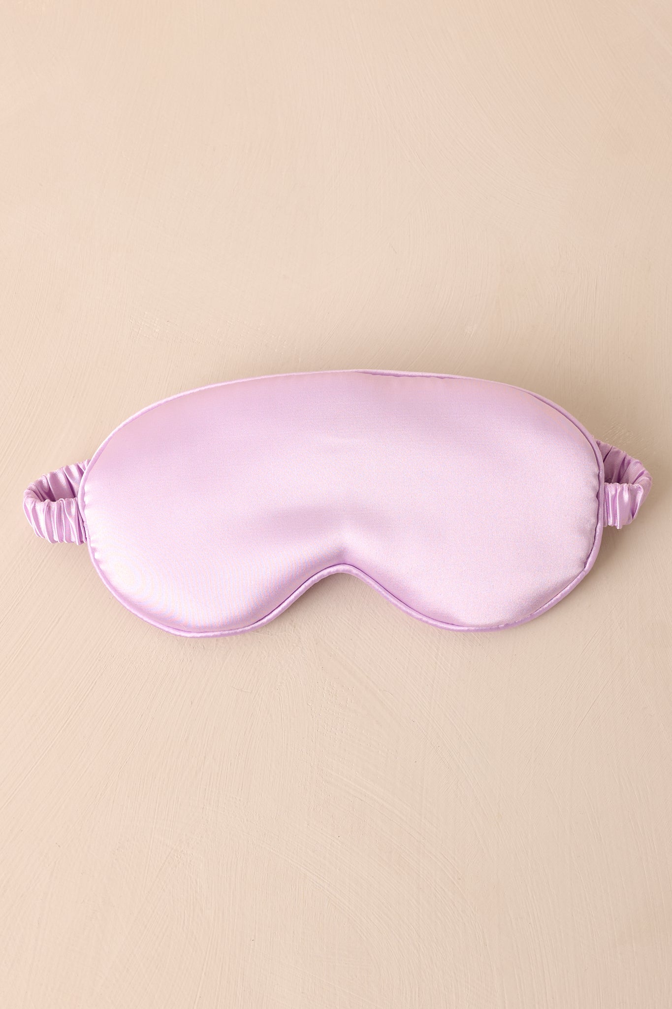 The lavender eye mask displayed against a neutral beige background, emphasizing its soft satin material and calming color.