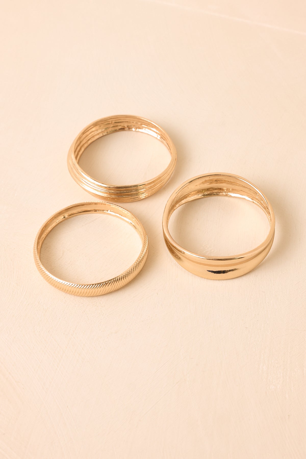 Layered gold bangles with polished, herringbone-inspired, and textured elements presented on a neutral backdrop.