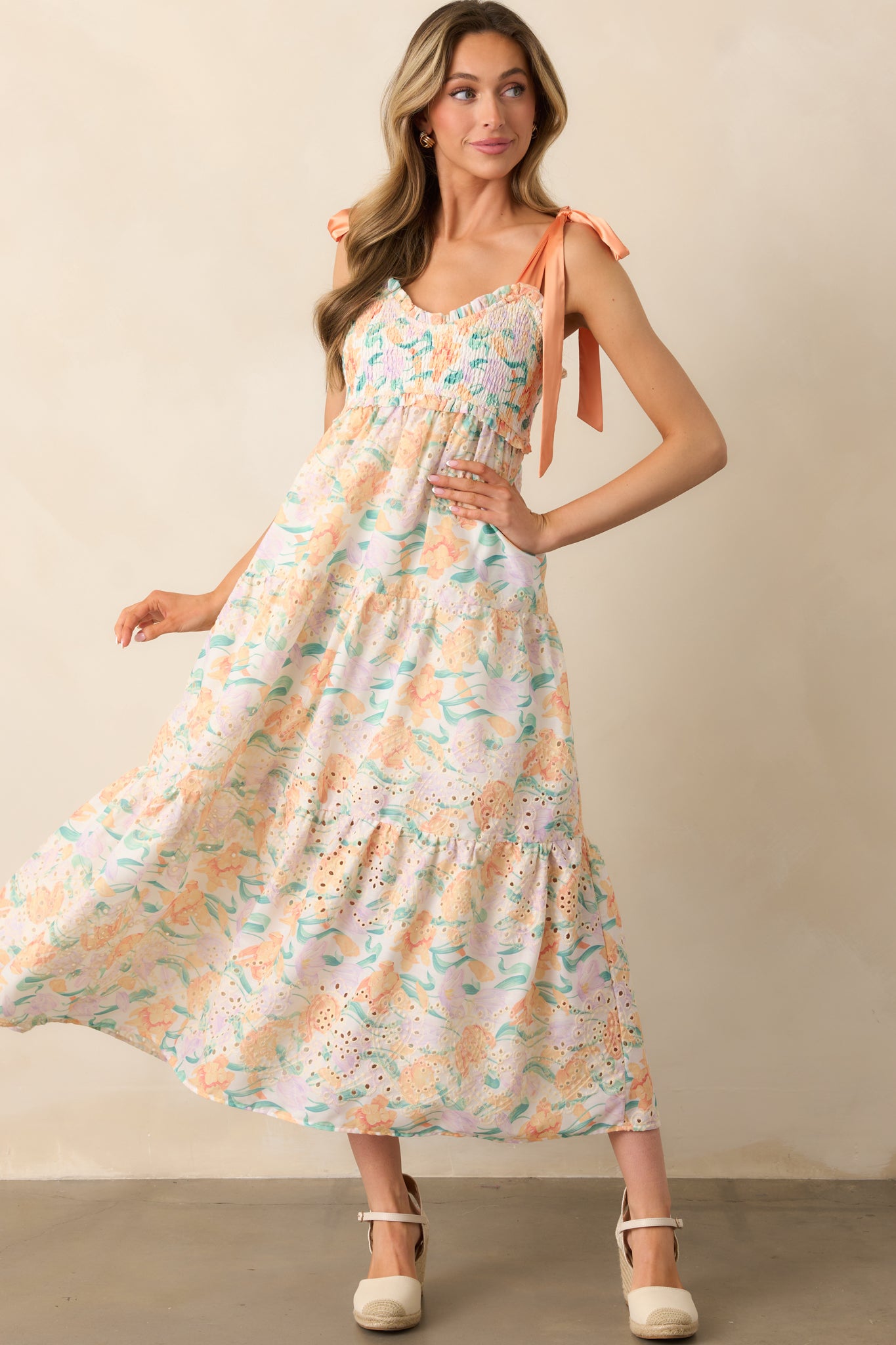 A light orange maxi dress featuring adjustable self-tie satin bow straps, a multicolor floral print, and a fully smocked bust for a flattering fit.