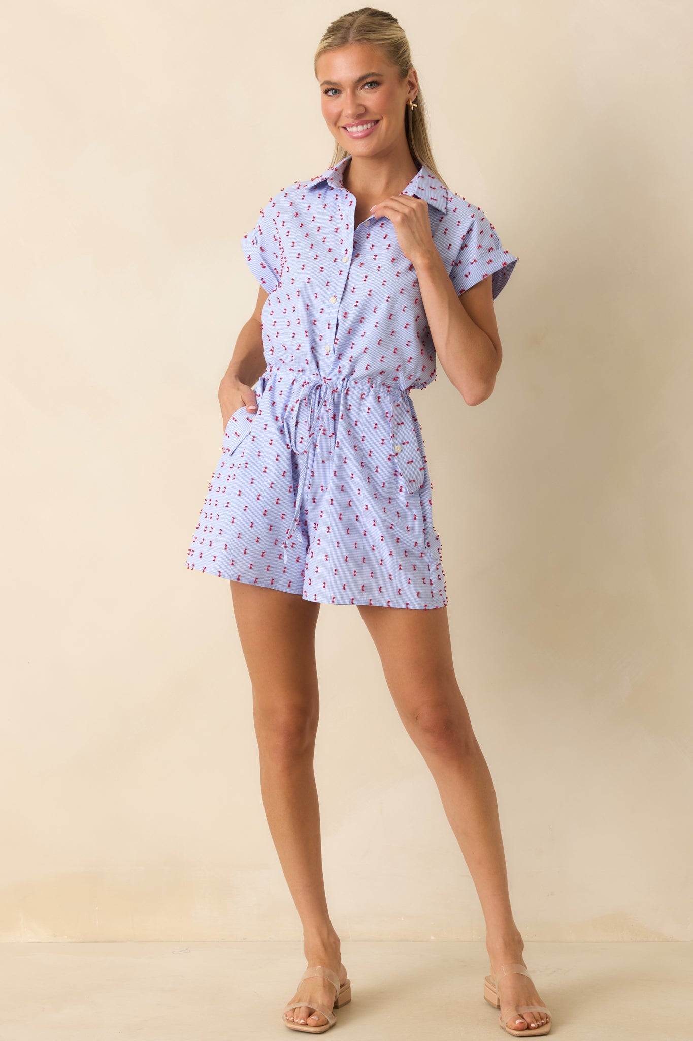 Lightweight blue gingham romper with a collared top, embroidered detailing, rolled sleeves, and an effortless warm-weather design.