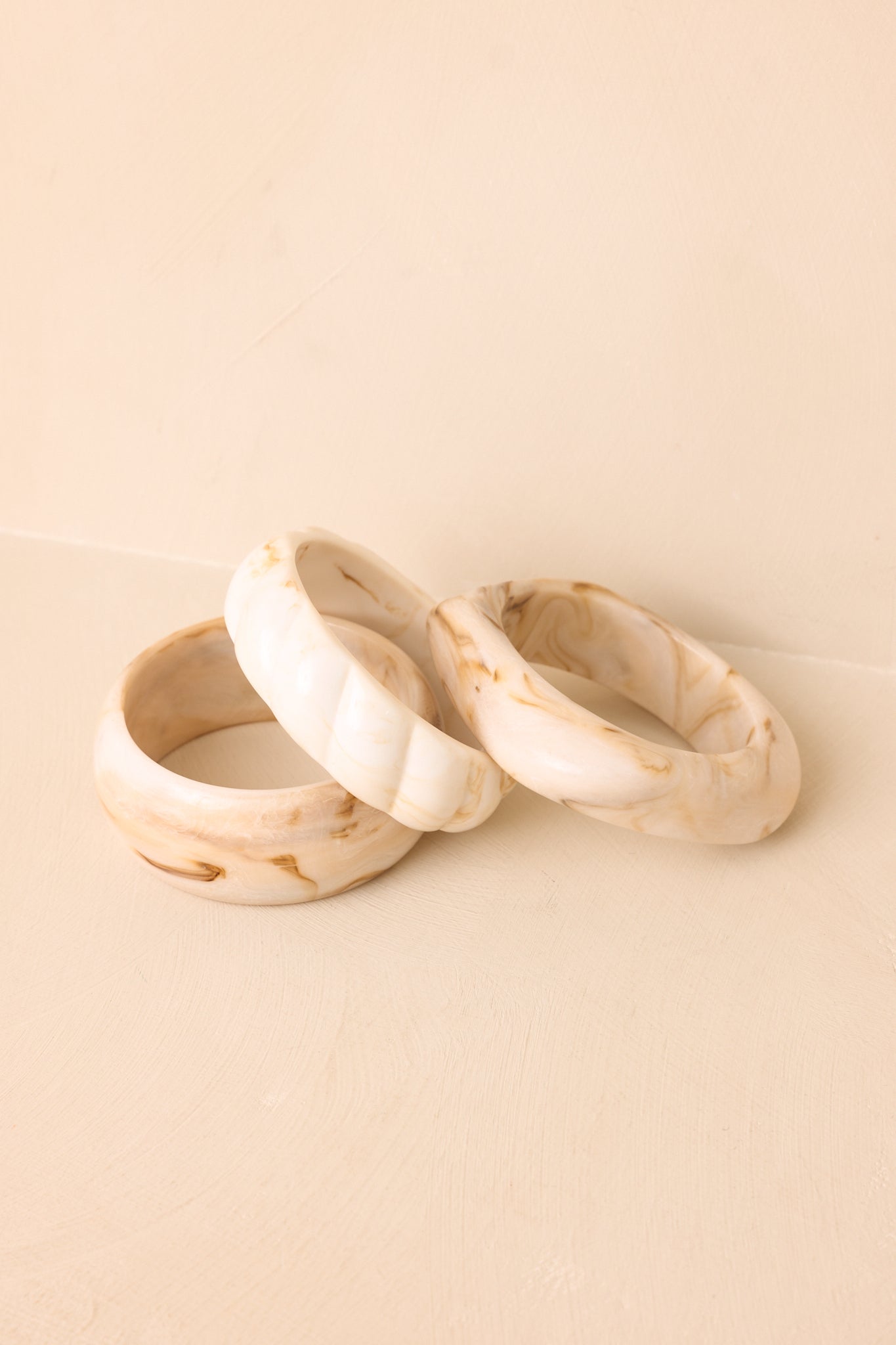 Trio of marbled ivory bangles with smooth sculptural designs displayed together against a beige background.