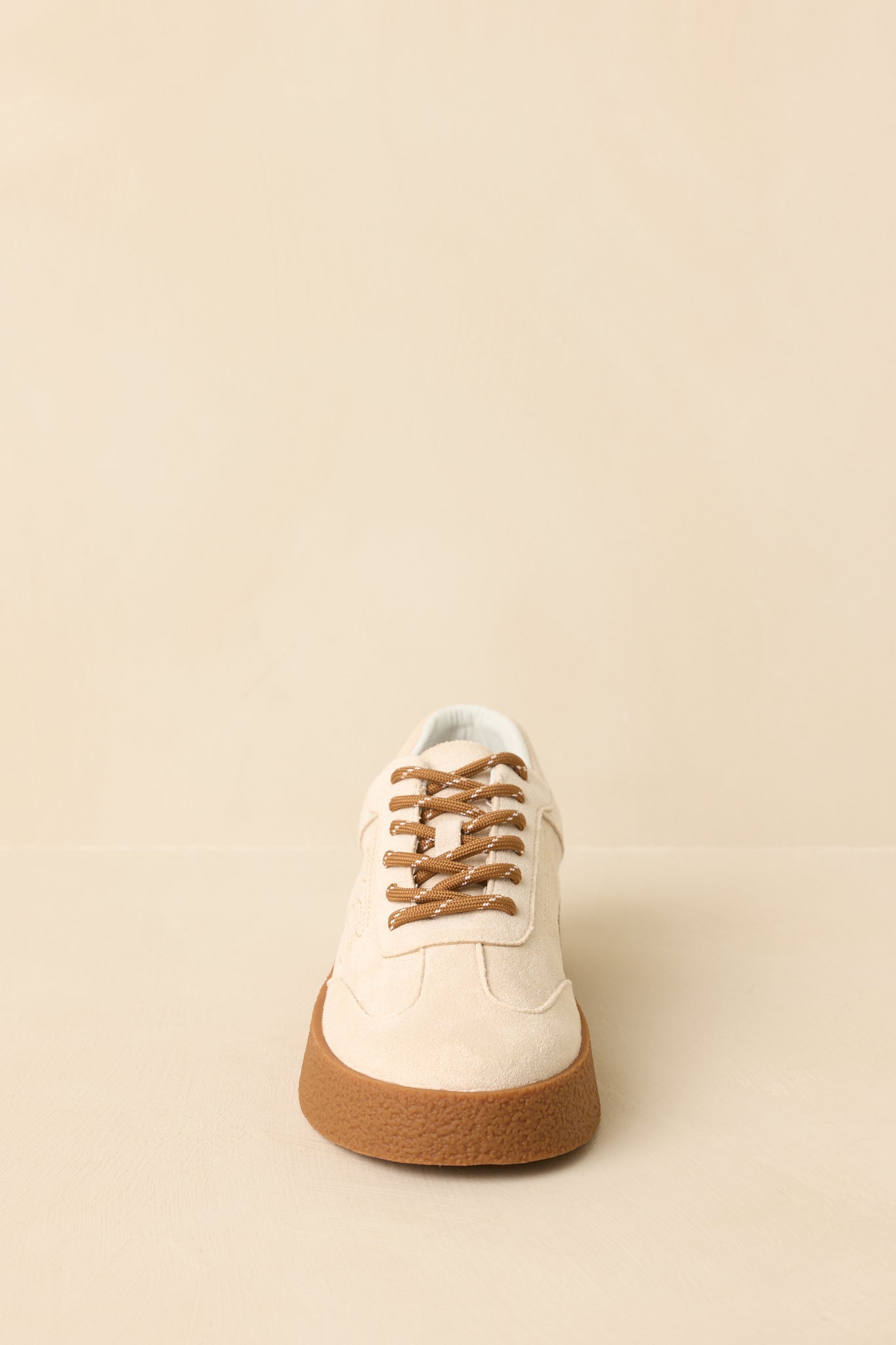 Front-facing view of natural faux suede sneakers against a beige background highlighting the rounded toe and lace-up design.