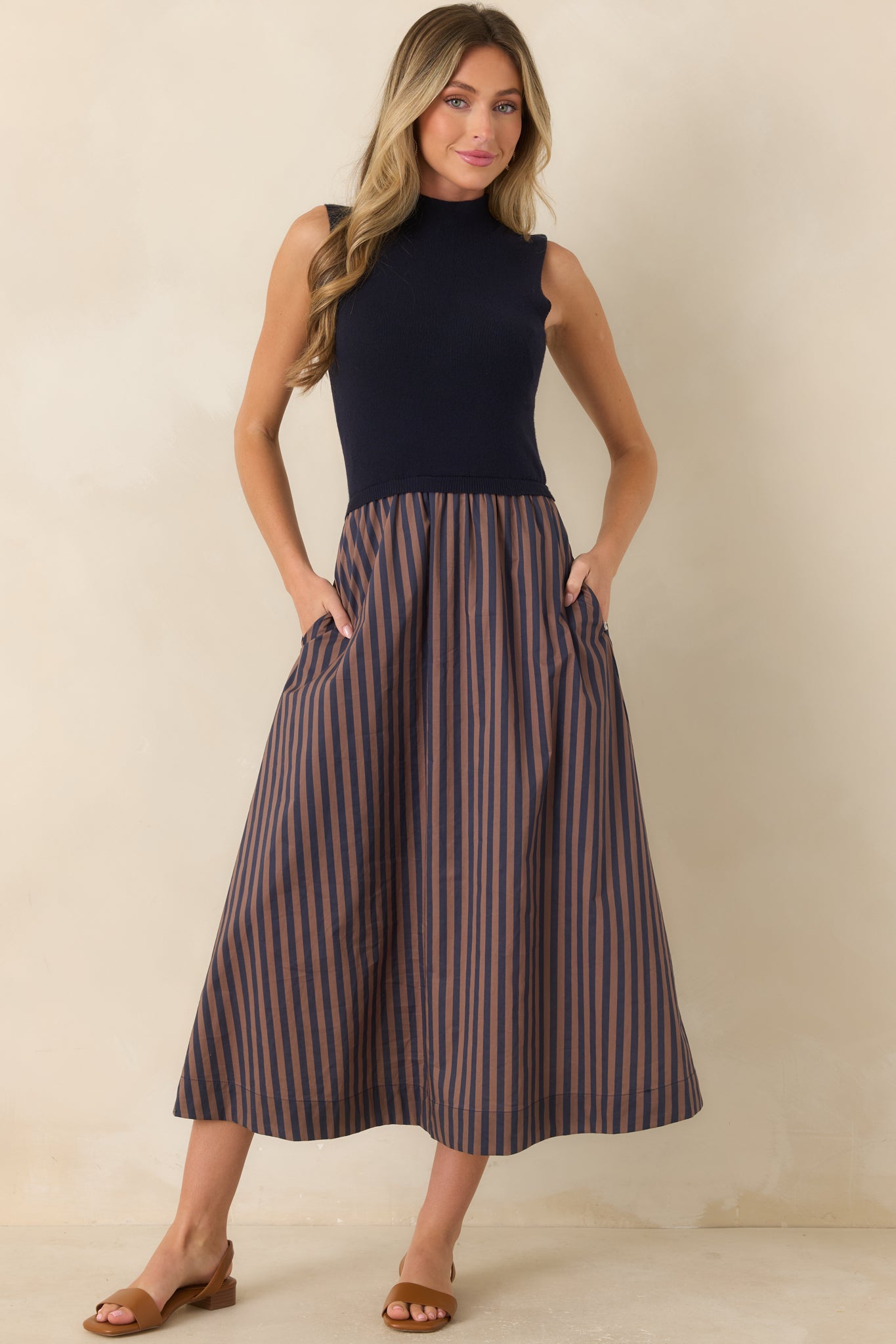 Navy sleeveless midi dress featuring a fitted bodice, elegant mock neckline, and a full skirt with warm tan stripes for a polished finish.