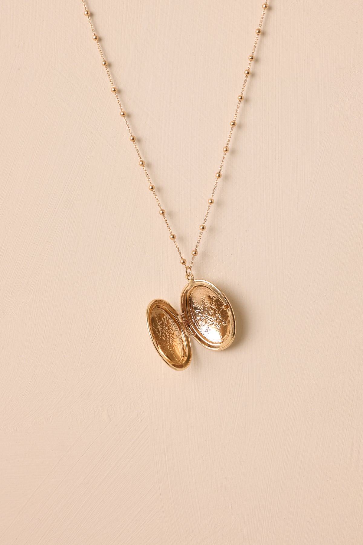 Angled view of the necklace laid flat against a beige background, emphasizing the size and subtle engravings on the oval locket.