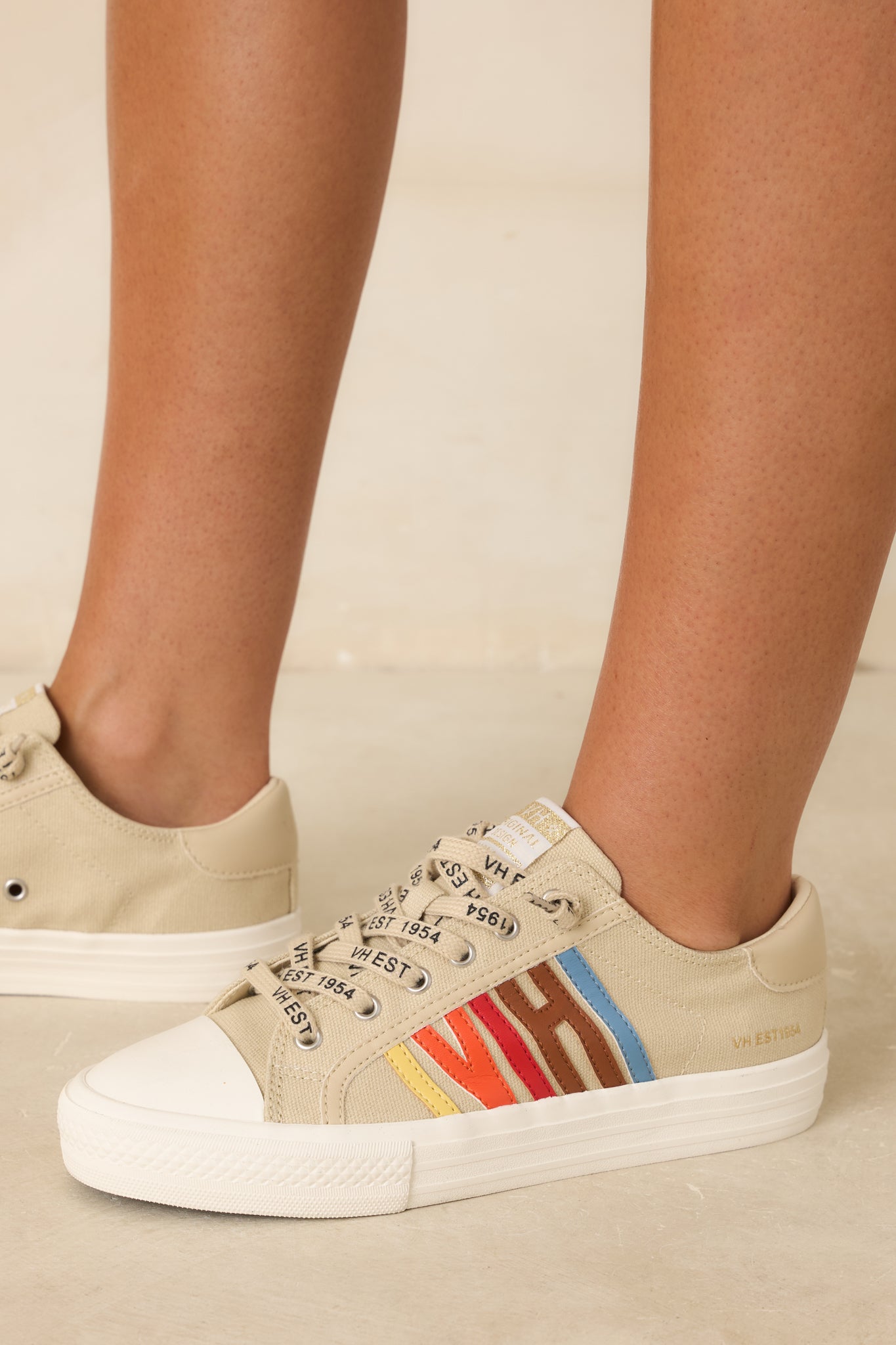 Nude canvas sneakers with multicolored side detailing and cushioned soles presented at a complementary angle against a soft beige backdrop.