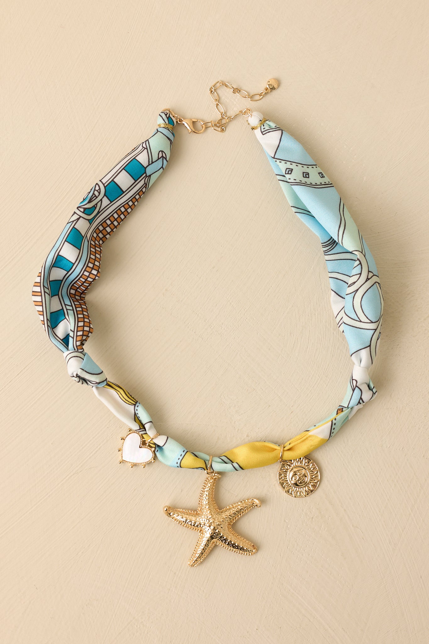 Ocean-inspired scarf necklace displayed against a beige background featuring blue tones and mixed gold charms.