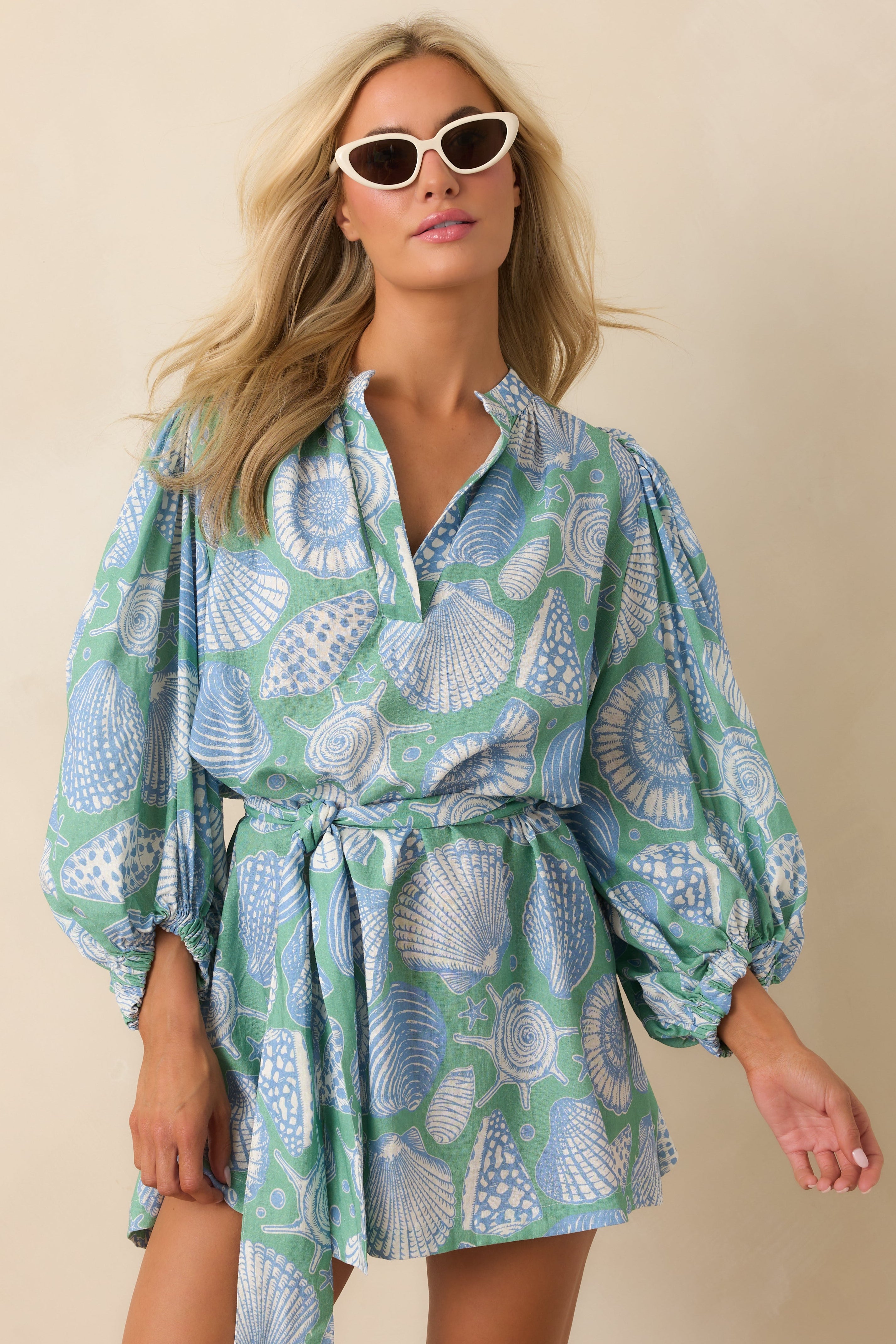Woman wearing a patterned dress with puff sleeves against a beige background. Dress is covered in sea shells.