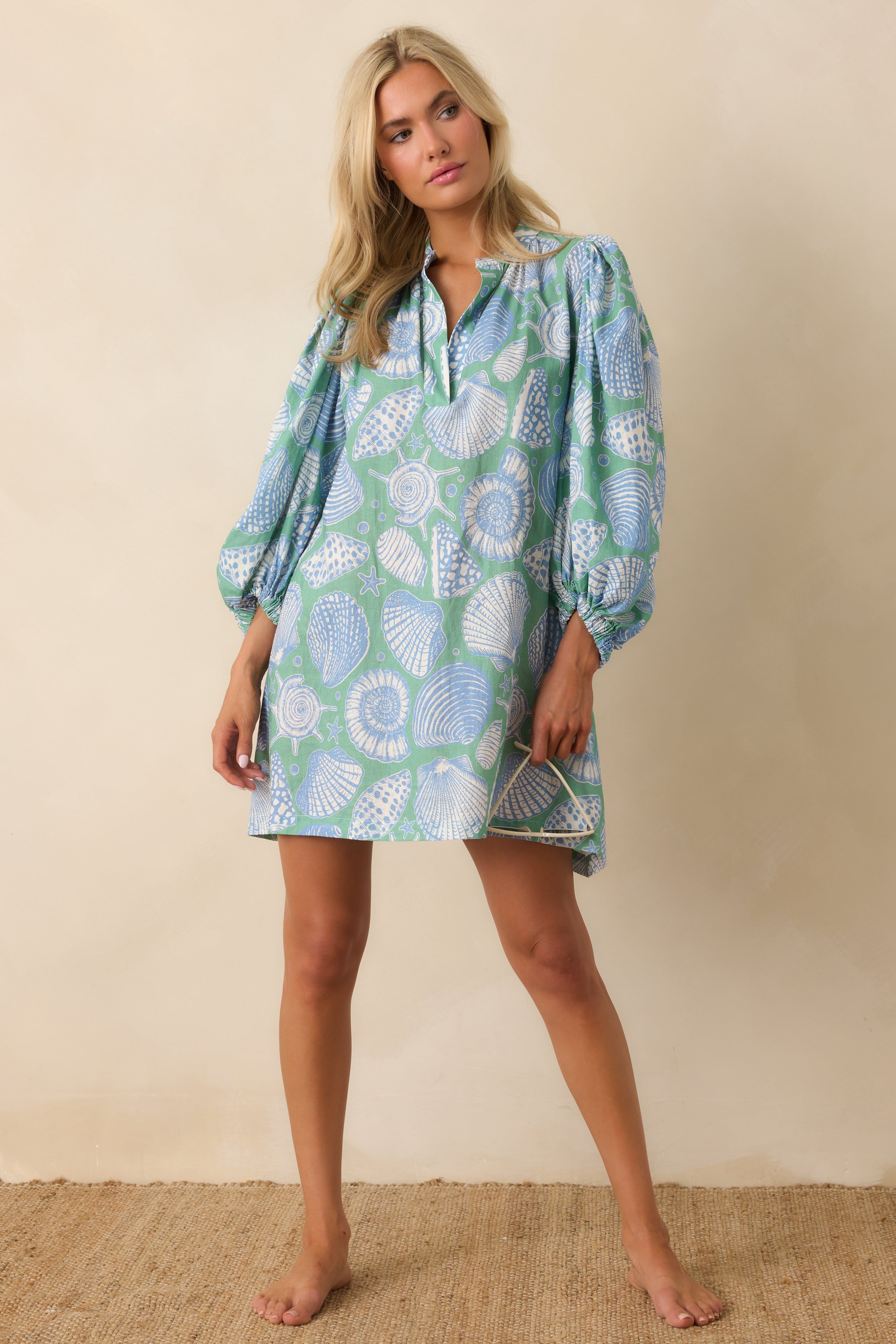 Woman wearing a blue and green patterned dress against a beige background. Dress has seashell print.