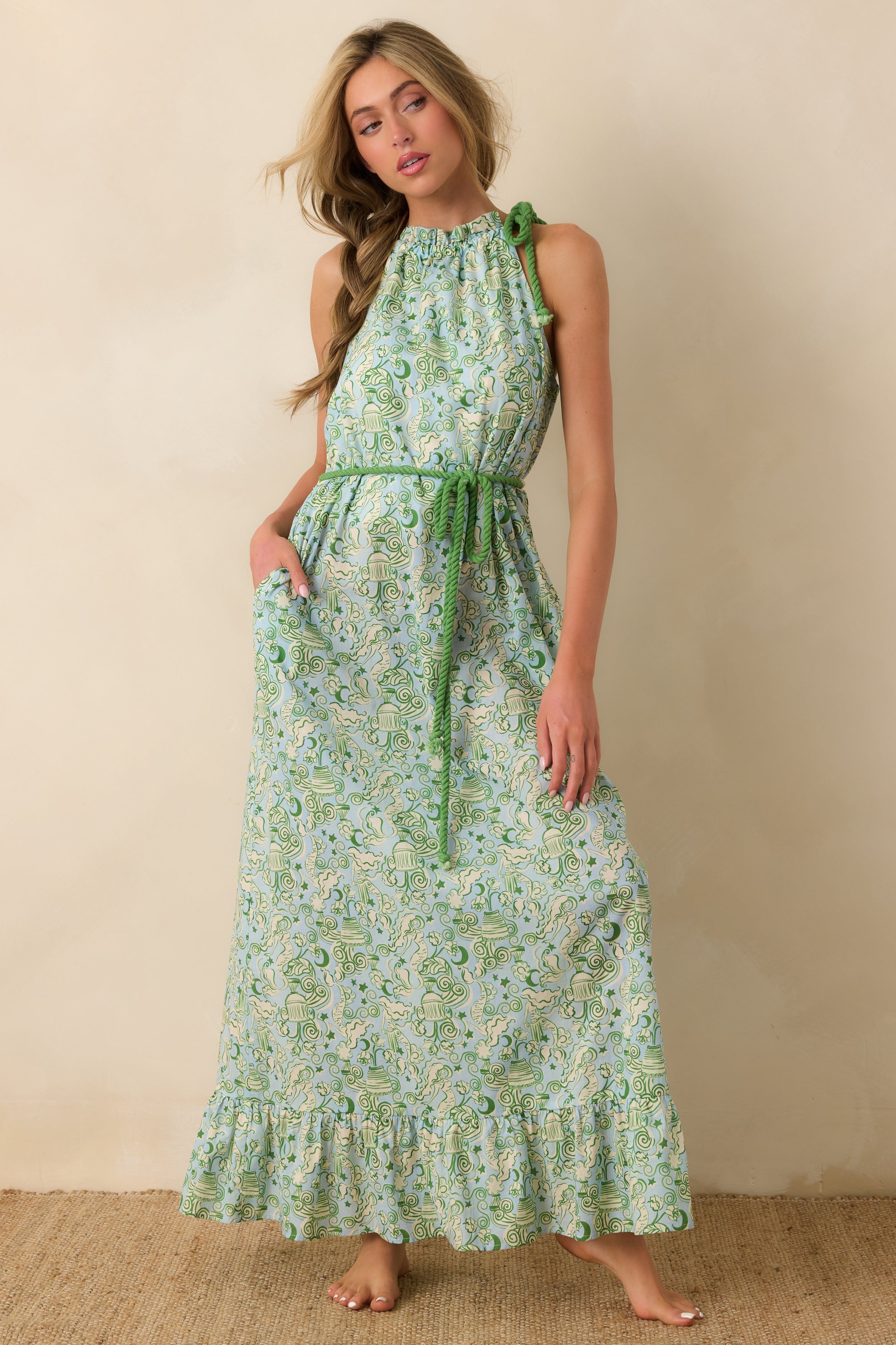 Woman wearing a green mermaid print maxi dress against a beige background. Dress has a green rope belt.