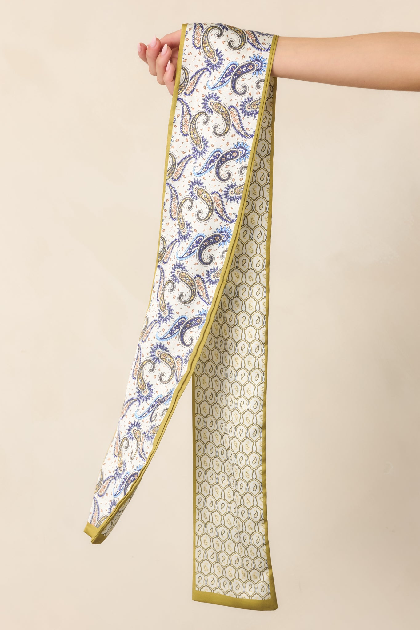 Close view of the hair scarf featuring its narrow shape, olive edging, and contrasting paisley motifs in earthy and blue tones.