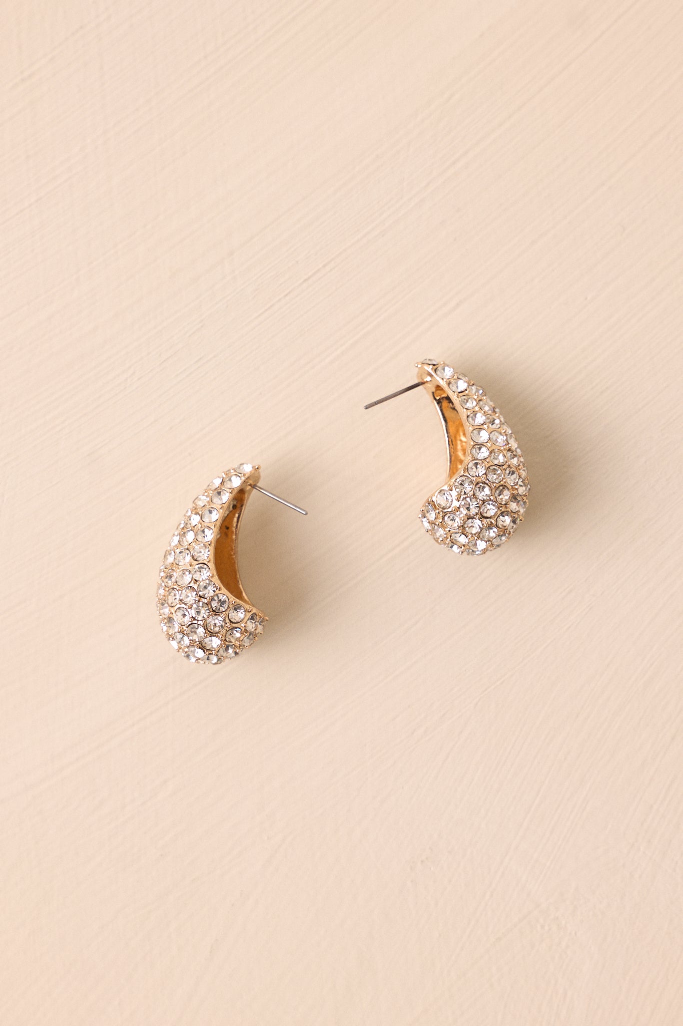 Pavé-style gold earrings with sparkling rhinestones and secure post backings shown on a beige backdrop.