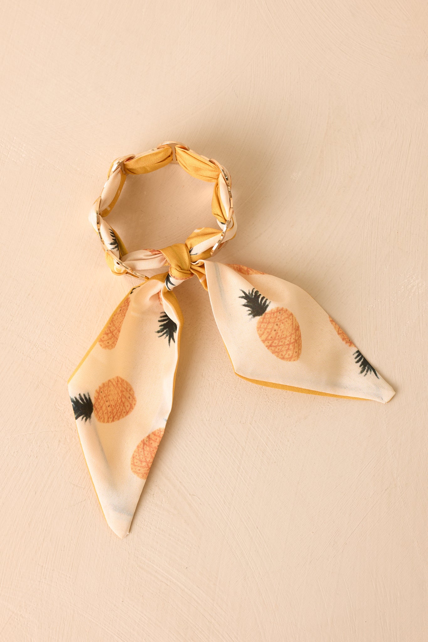 Pineapple-print scarf woven through a gold cuff bracelet photographed against a beige background.